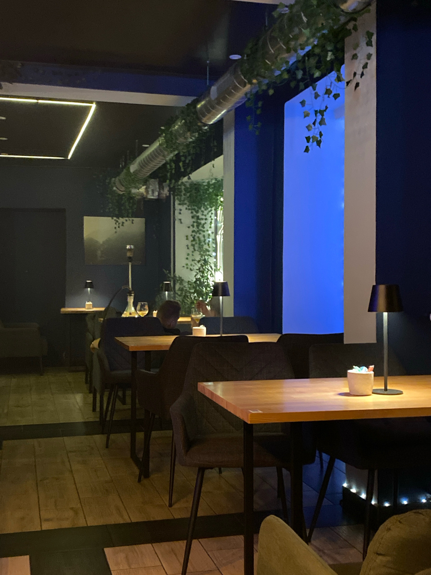 The image depicts the interior of a dimly lit, modern establishment, likely a hookah lounge or bar, with a dark color palette dominated by blues and blacks, accented by warm wooden furniture and strategically placed lighting. The space appears to be located in Gliwice, Poland, suggesting a contemporary urban setting.
In the foreground, a light wooden table with black legs is set with a small white ceramic pot containing colorful objects, possibly candy or decorative items, and a sleek black table lamp. A person's shoulder and head are visible in the lower right corner, leaning back on a plush seat.
Moving further into the scene, several more identical tables and dark upholstered chairs are arranged in rows, suggesting a seating area designed for multiple patrons. A hookah, a tall apparatus with a glass base and a metal stem, stands on a table in the mid-ground. Two wine glasses are also visible on this table, one partially filled.
A person, a young male with dark hair, is seated at the table with the hookah, facing away from the camera and slightly turned to the side, appearing to be engaged with something on the table.
The walls are painted a deep navy blue, with one section featuring a large, illuminated rectangular panel emitting a bright, cool blue light. This creates a striking contrast with the darker areas of the room.
Decorative elements include artificial ivy vines cascading from a large, metallic ventilation pipe that runs horizontally across the ceiling. A framed abstract artwork hangs on the dark wall to the left. The floor is covered in wooden planks with dark linear inlays. The overall ambiance is one of relaxed sophistication, with a focus on mood lighting and modern design.