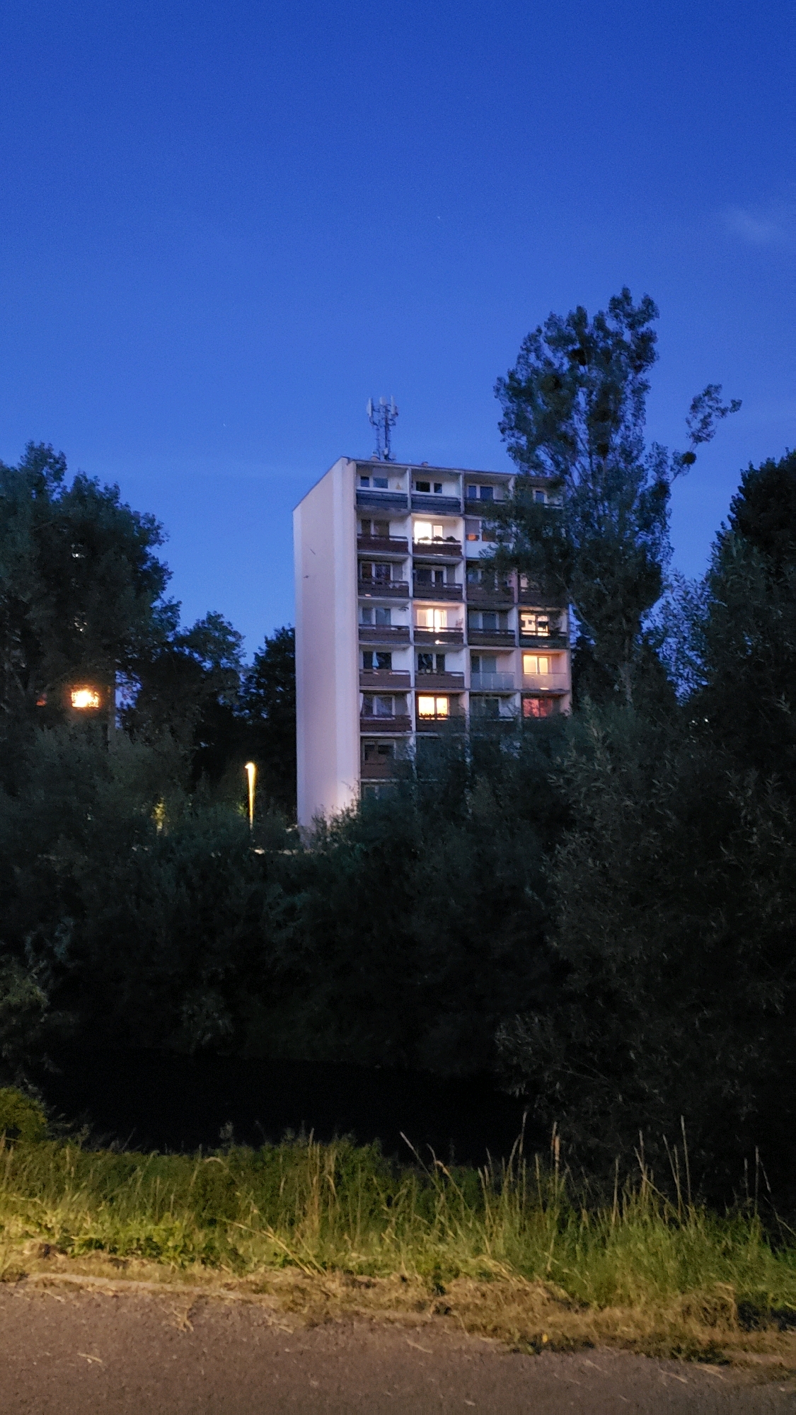 An exterior, vertical, low-angle shot captures a tall apartment building at dusk in Prešov, Slovakia. The building, with a light-colored facade, has multiple floors featuring balconies with railings and numerous windows, some of which are illuminated with warm, yellow light, suggesting occupancy. A telecommunications antenna is visible on the rooftop. The apartment building is partially obscured by dense, dark green foliage from trees and bushes in the foreground and mid-ground. To the left of the building, a separate light source, possibly a streetlamp, is visible within the trees, emitting a warm glow. In the foreground, a strip of tall, dry grass runs along the edge of what appears to be a paved surface, possibly a road or sidewalk. The sky above is a deep, clear blue, transitioning into twilight.