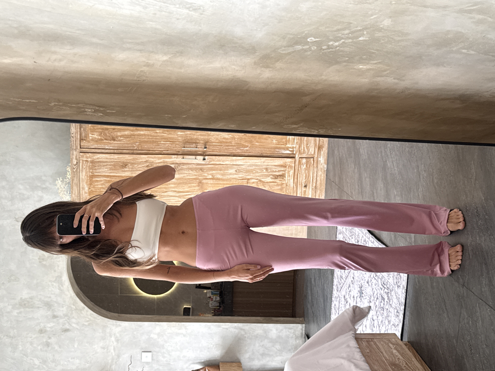 A woman is standing barefoot in front of a mirror, holding a smartphone in her left hand, presumably taking a picture of herself. She is wearing a white top and mauve-colored yoga pants. Her brown hair is wavy. The reflection shows a room in what appears to be a residence in North Kuta, Indonesia, judging by the style of the decor and architecture. A wooden dresser is visible in the mirror, as well as a partially visible bed with white linens, and gray tiling on the floor. A doorway with a curved arch reveals what looks like a bathroom area with a built-in light fixture.