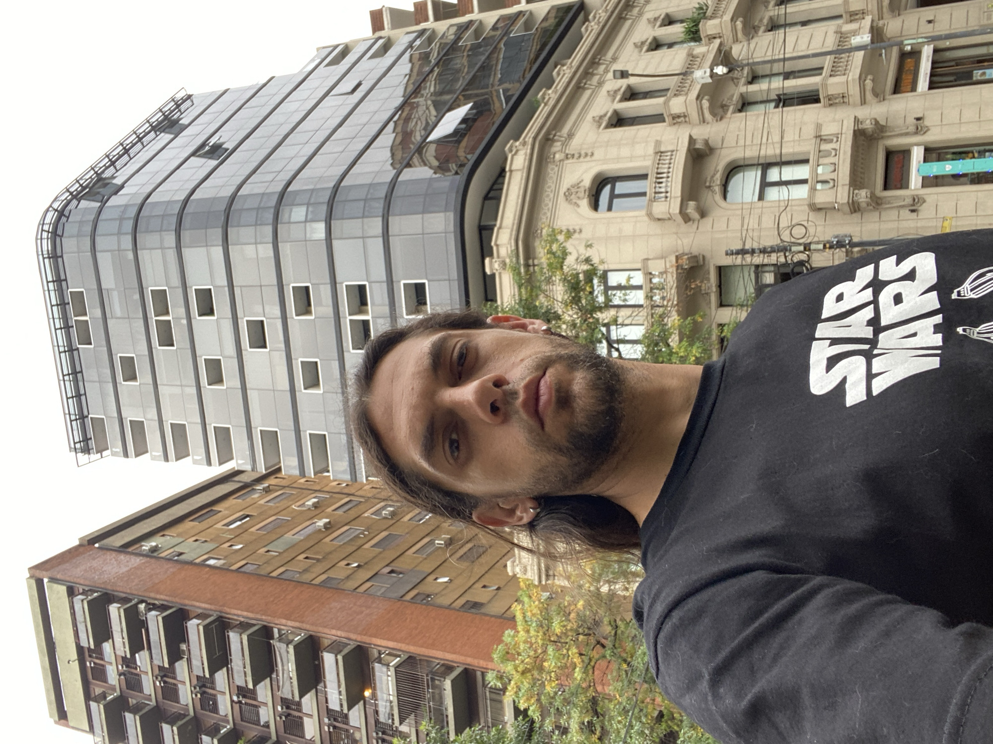 A male individual is featured in a close-up, eye-level selfie taken in the city of Buenos Aires, Argentina, specifically near San Martín 683. He has long dark hair, a beard, and a neutral expression, looking directly into the camera. He is wearing a black t-shirt with "STAR WARS" printed in white capital letters across the chest. A silver hoop earring is visible in his left ear. The background showcases an urban environment with three distinct architectural styles. To the subject's right, a modern multi-story building with a curved, dark glass facade and integrated light-colored window units rises. Below this, and extending to the right, is a classical-style, light beige building with ornate detailing, balconies with balustrades, and arched windows, situated at a corner. To the subject's left, an older, tall residential building with a brown brick facade and numerous balconies is visible. Green and yellow-leafed trees are present between the subject and the background buildings. The sky above is overcast and light gray.