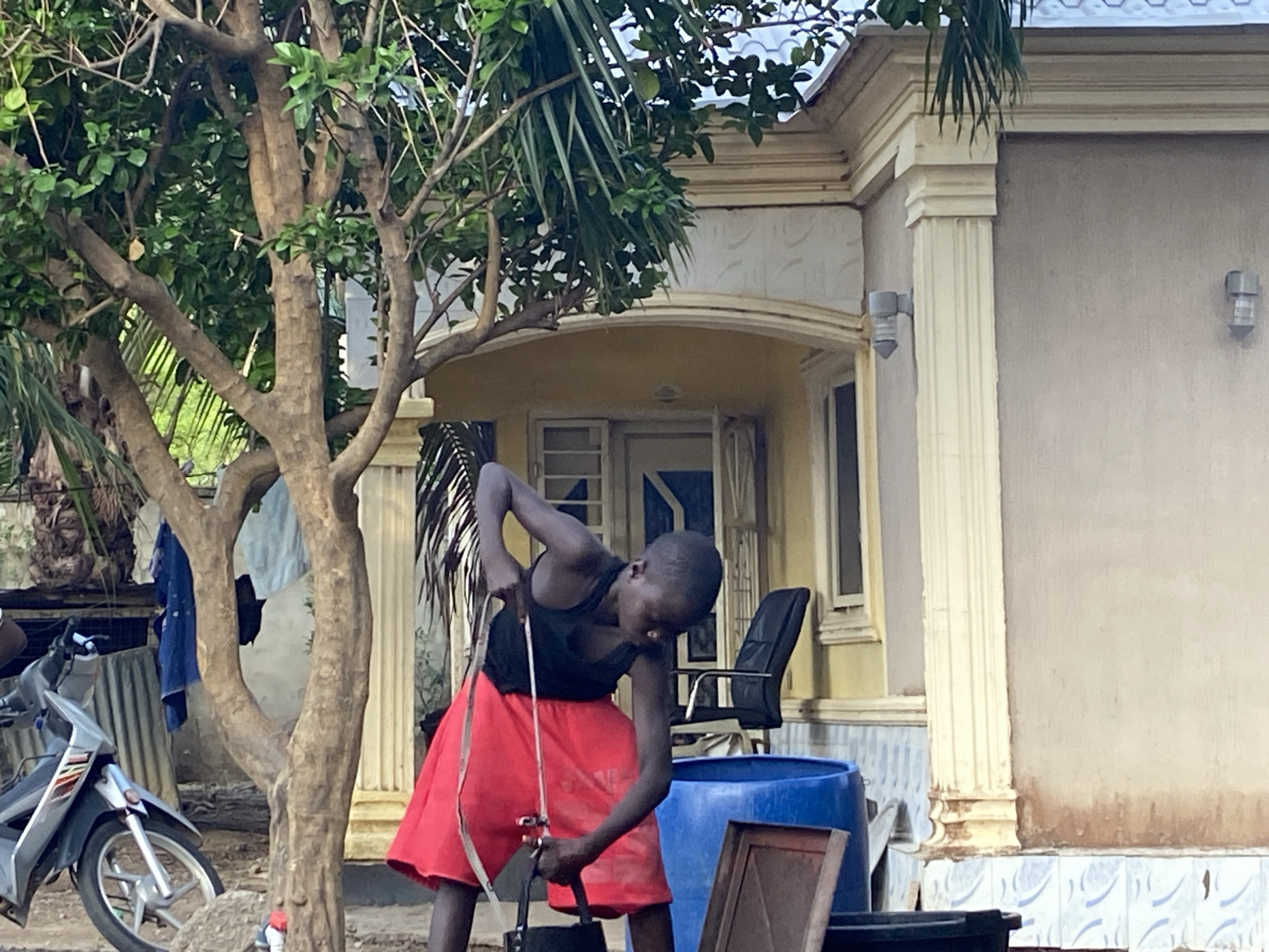 A young individual, likely a child, wearing a black tank top and a red skirt is working outside a building with a cream-colored facade. The individual is bent over and appears to be working with some containers, possibly transferring liquids between them. A blue plastic barrel and other dark-colored containers are present.
In the background, a house is partially visible. It features a covered entrance supported by columns, and the architecture suggests a residential structure. A black office chair is visible through the open doorway.
To the left, a tree with thin branches and green leaves frames the scene. A motorbike is parked to the left of the tree, and some laundry is hanging on the tree. The location is likely in Mararaba, Nigeria.