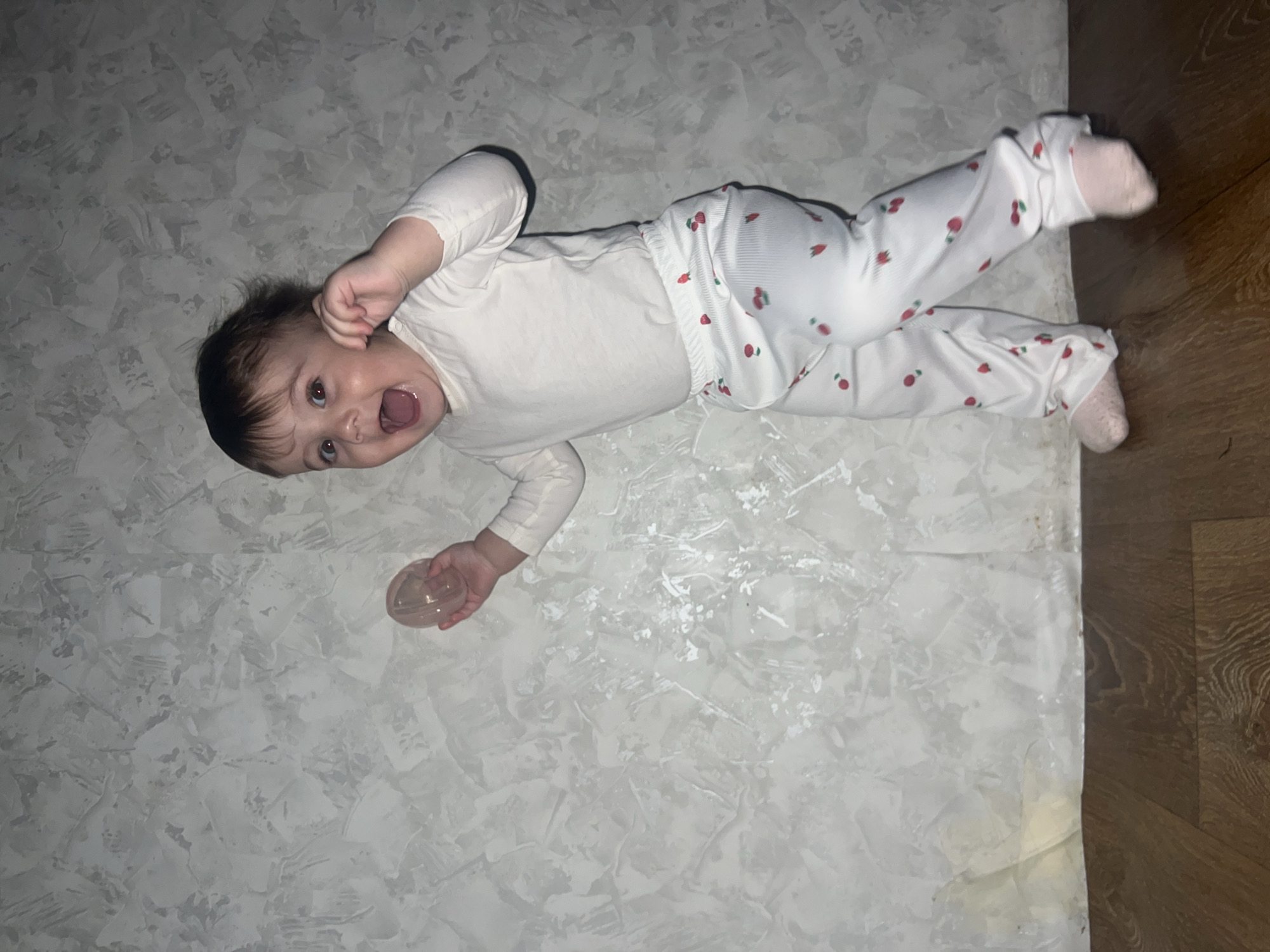 A baby in white pajamas with red cherry designs is lying on a gray and white patterned surface. The baby's mouth is open, and they appear to be shouting. They are holding a clear plastic object in their left hand. The edge of the patterned surface meets a wooden floor on the right side of the image. The location is unknown, and there is no indication of the city of Elena, Bulgaria, in the image.