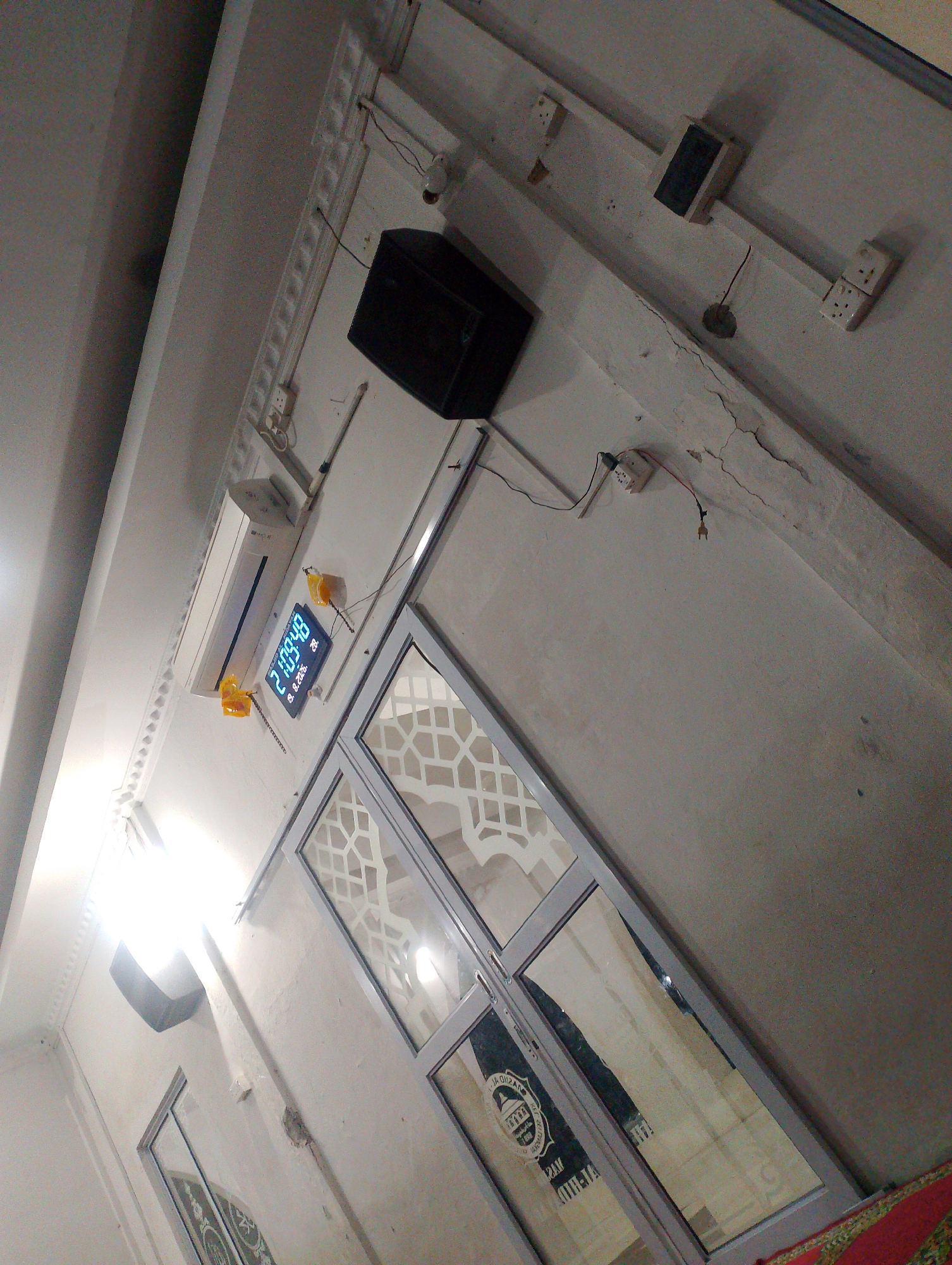 The image depicts the interior of a building, likely a mosque, given the presence of Islamic calligraphy on a glass door. The perspective is from a low angle, looking upwards towards the ceiling. Prominently displayed on the wall is a digital clock showing "21:09:48", along with a date that appears to be "26.10.2006". Above the clock is a white air conditioning unit. To the left of the clock are two small yellow objects with chains, possibly decorative elements or part of the clock's mechanism. A black speaker is mounted high on the wall near the ceiling. Electrical outlets and wiring are visible along the wall and ceiling. The walls appear to be painted white and show some signs of age, including cracks. A glass door with decorative grilles is visible in the lower right portion of the image. The presence of a digital clock with the date suggests the image was taken on October 26, 2006. Without further information or context, it is not possible to definitively determine the specific location within Batam, Indonesia.