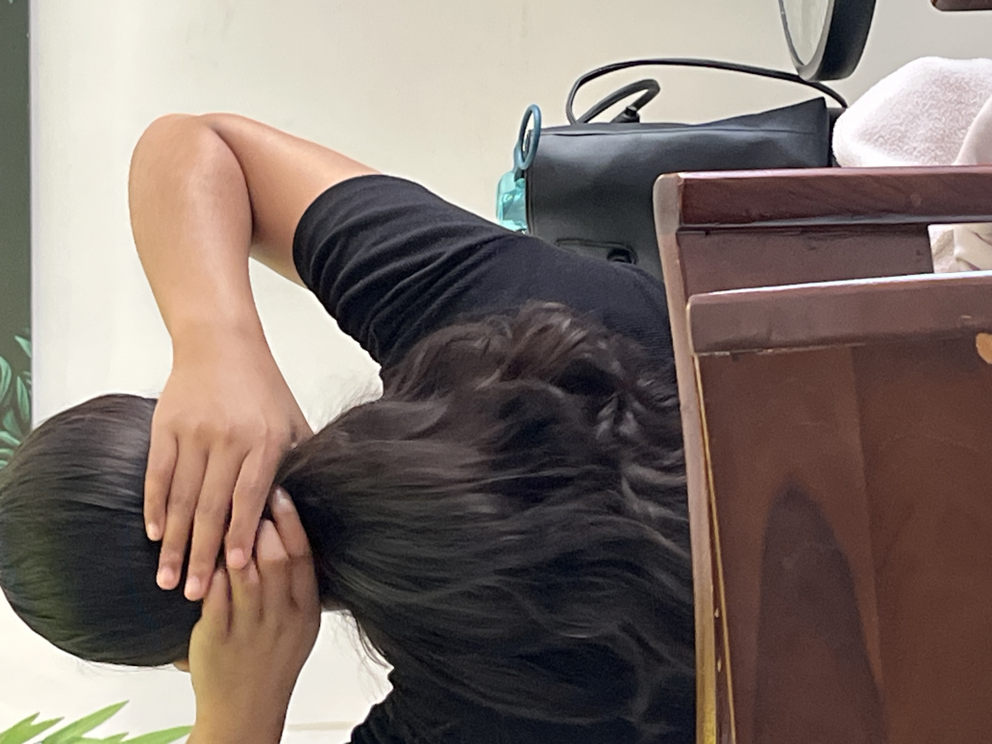 The photograph shows a person with dark hair, wearing a black shirt. They are holding their hair up with both hands, possibly styling or adjusting it. In the background, a black bag is visible, along with what appears to be a white towel and a wooden chair. A water bottle is also partially visible. The location is Unknown, Indonesia.