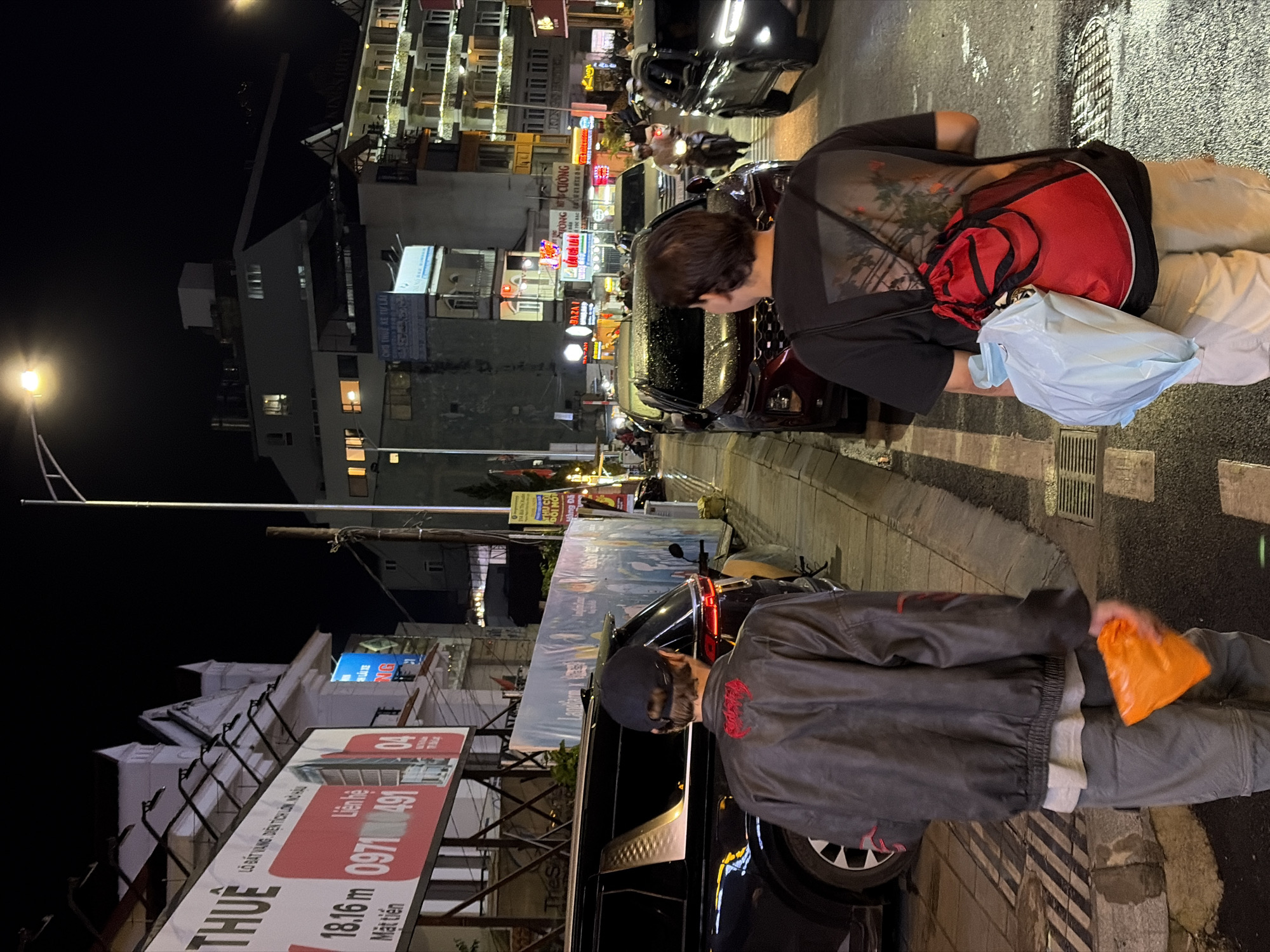 This is a nighttime, eye-level, medium shot of two young men walking away from the camera on a street in Da Lat, Vietnam. The street is wet, suggesting recent rain, and illuminated by streetlights and the lights from the surrounding buildings. Both men are wearing casual clothing. The man on the left is wearing a black bomber jacket with red accents and grey pants. He is holding a bright orange bag in his right hand. The man on the right is wearing a black t-shirt with a graphic print on the back and light-colored cargo pants. He has a red drawstring bag slung over his shoulder, and a light blue plastic bag is tucked under his arm.
On the left side of the street, there are several parked vehicles, including a black SUV. A large billboard with Vietnamese text and images is mounted on the building behind the SUV. The building itself appears to be a commercial establishment, with signs and architectural features indicative of the city's European-influenced style.
On the right side of the street, more cars are parked, and a busy intersection is visible in the background with traffic, including motorcycles. The buildings on this side are multi-story structures with illuminated windows and balconies, typical of the urban landscape in Da Lat. The overall atmosphere is lively and urban, despite the late hour and wet conditions.