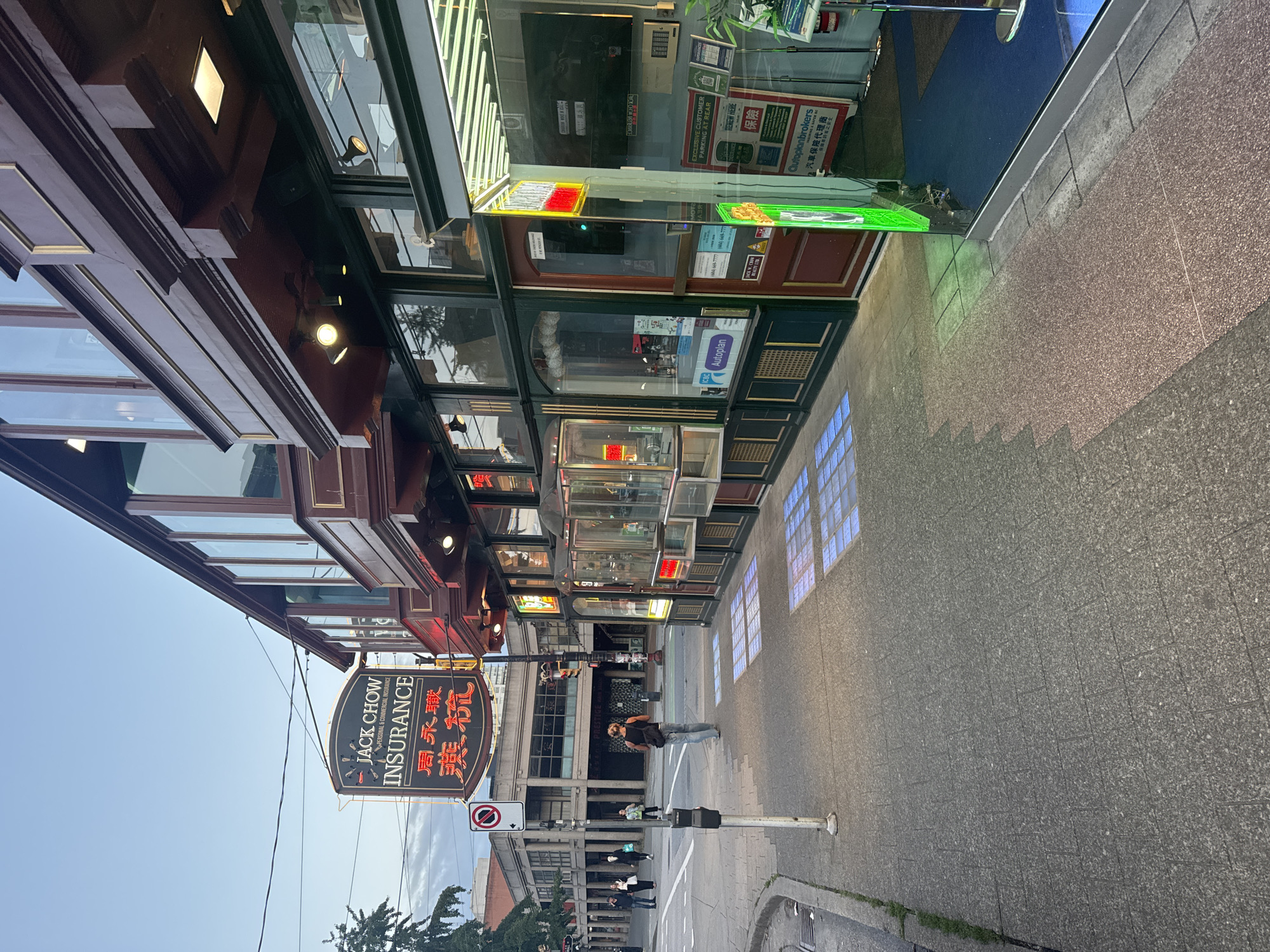 The image shows a street scene in Vancouver, Canada. In the foreground, a sidewalk is paved with concrete pavers. To the right, the storefront of "Jack Chow Insurance" is visible, featuring a neon sign with English and Chinese characters. The building has a dark wood facade and large glass windows displaying insurance-related posters and advertisements. A red door is also present.
On the sidewalk, a person wearing a dark tank top and jeans walks away from the camera. In the background, other pedestrians are seen walking on the sidewalk. Across the street, a building with a concrete facade and multiple columns is visible. A traffic light and a "No Parking" sign are also present. The sky is overcast with a pale blue hue. The overall scene suggests an urban environment, possibly a commercial district.