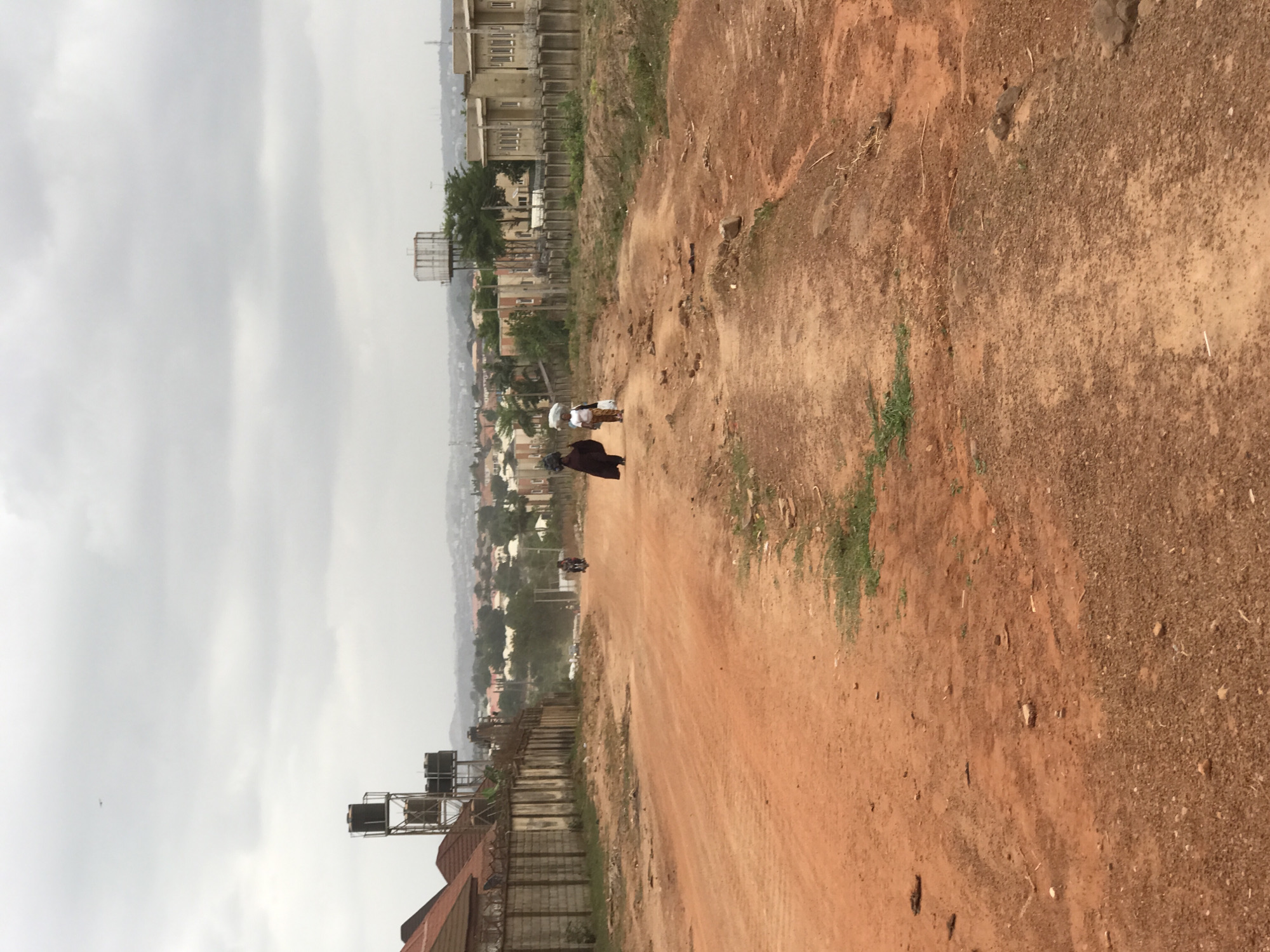 The image depicts a dirt road in Abuja, Nigeria. The road is predominantly reddish-brown and appears uneven. Two individuals are walking along the road: one in a brown robe and one wearing a white top. The environment includes buildings and vegetation in the background. The sky is overcast. Water tanks on a rooftop are visible in the foreground.