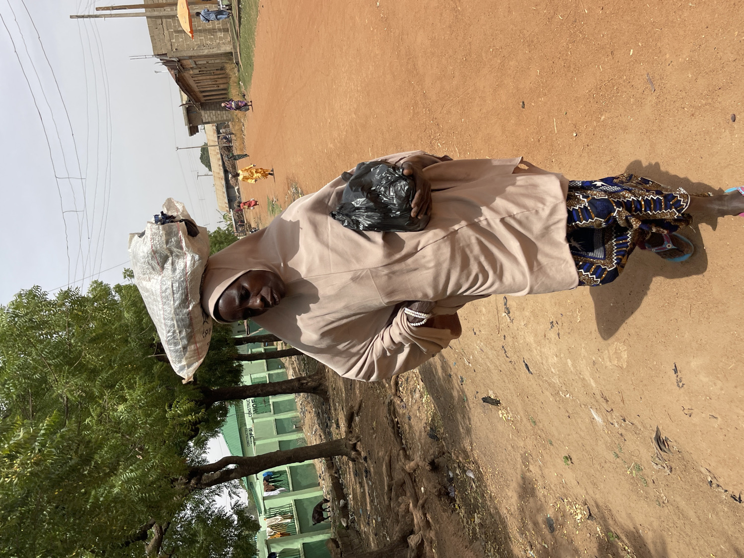 This media file depicts a person, likely a woman, walking or standing on a reddish-brown dirt road in Muchiya, Nigeria, under bright daylight. The subject is seen from the waist up, wearing a light-colored (beige or light brown) headscarf and a matching long-sleeved outer garment. A patterned dark blue and brown skirt or wrapper is visible below. On her right wrist, she wears a light-colored beaded bracelet, and she has blue and brown sandals on her feet.
She is carrying a large, bulky, light-colored sack balanced on her head, and a smaller black plastic bag is held in her left hand, clutched to her chest. Her expression is neutral as she looks slightly to her left, towards the viewer.
The setting is an outdoor, possibly semi-urban or rural environment. To the left, several green-leafed trees are visible. In the mid-ground, behind the subject, there are single-story buildings, some appearing light green, along with utility poles and overhead power lines. On one of the light green buildings, partially visible text reads "CONSTRUCTED OFPAI". Further down the dirt road, other distant figures are seen, including someone in a bright yellow garment. The bright sunlight casts distinct shadows on the ground, indicating a clear, warm day. The scene portrays a moment of daily life, likely involving the transport of goods.