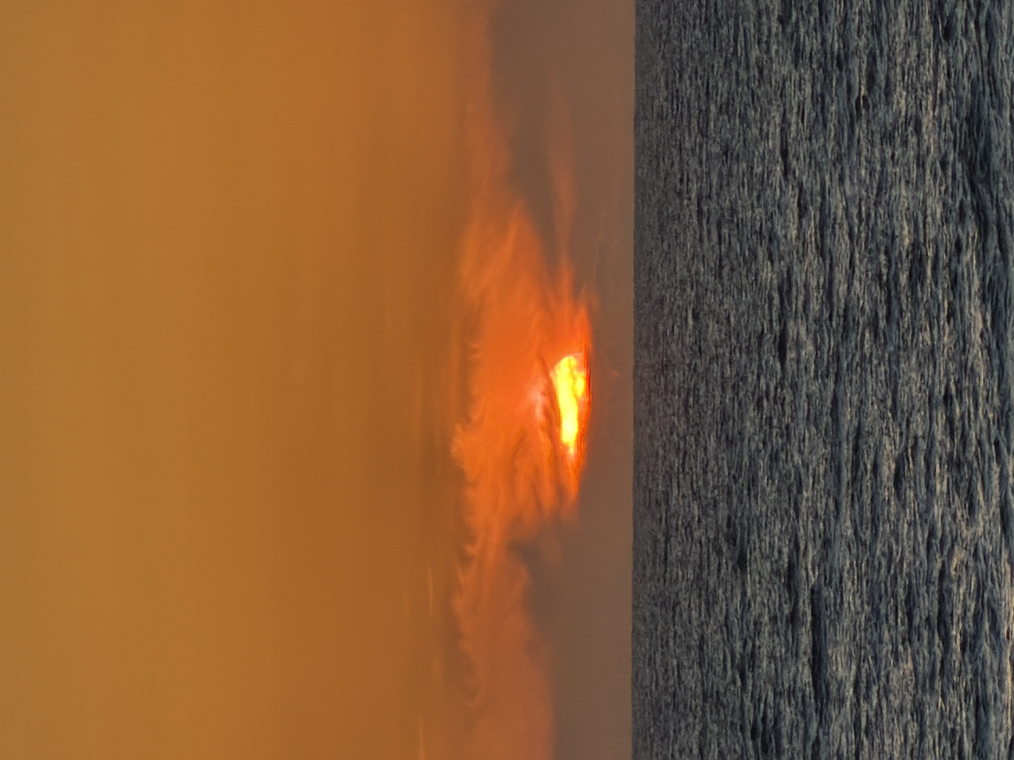 The image shows a hazy, orange sky on the left and a textured, dark-colored wall or rock face on the right. There's a bright, undefined light source obscured by haze or smoke in the middle of the left side. The location and details of people, subjects, objects, actions, interactions, and the environment are not discernible beyond what is described. The location in Tel-Aviv, Israel, cannot be confirmed or substantiated based on the visual information provided.