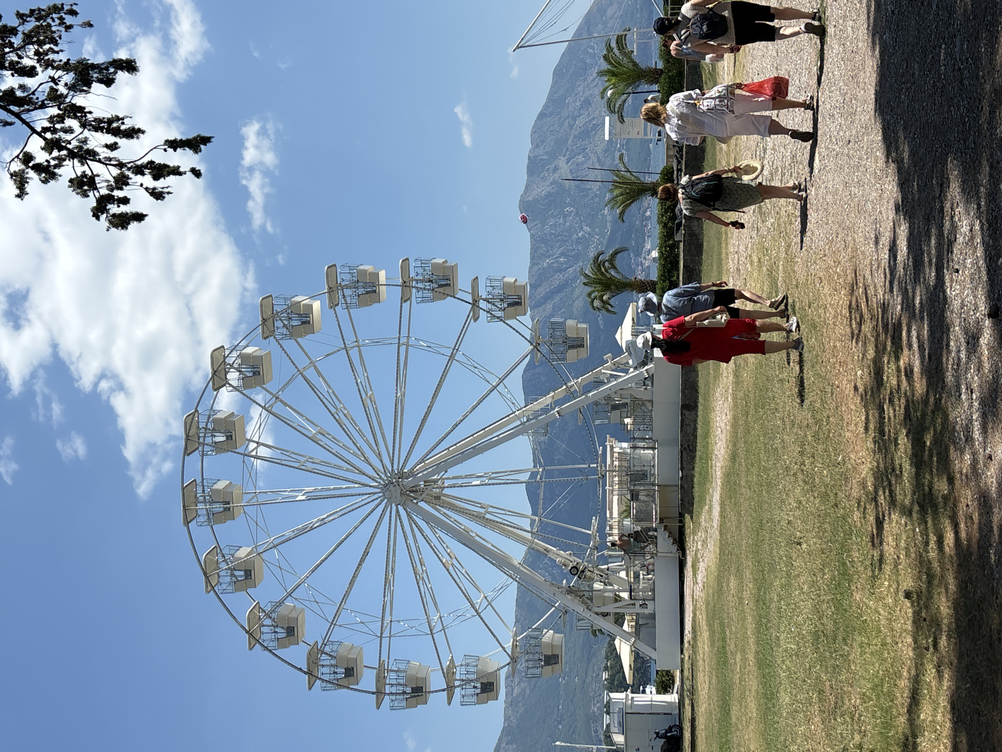 In Budva, Montenegro, a Ferris wheel stands tall against a clear blue sky dotted with white clouds. The Ferris wheel is white and has many passenger cabins. In the foreground, a group of people are walking away from the camera on a dirt path. Some are carrying bags and one person is wearing a hat. The ground is covered in grass and shadows of trees. In the background, there are palm trees, mountains, and the sea. A red object is visible in the sky above the mountains.