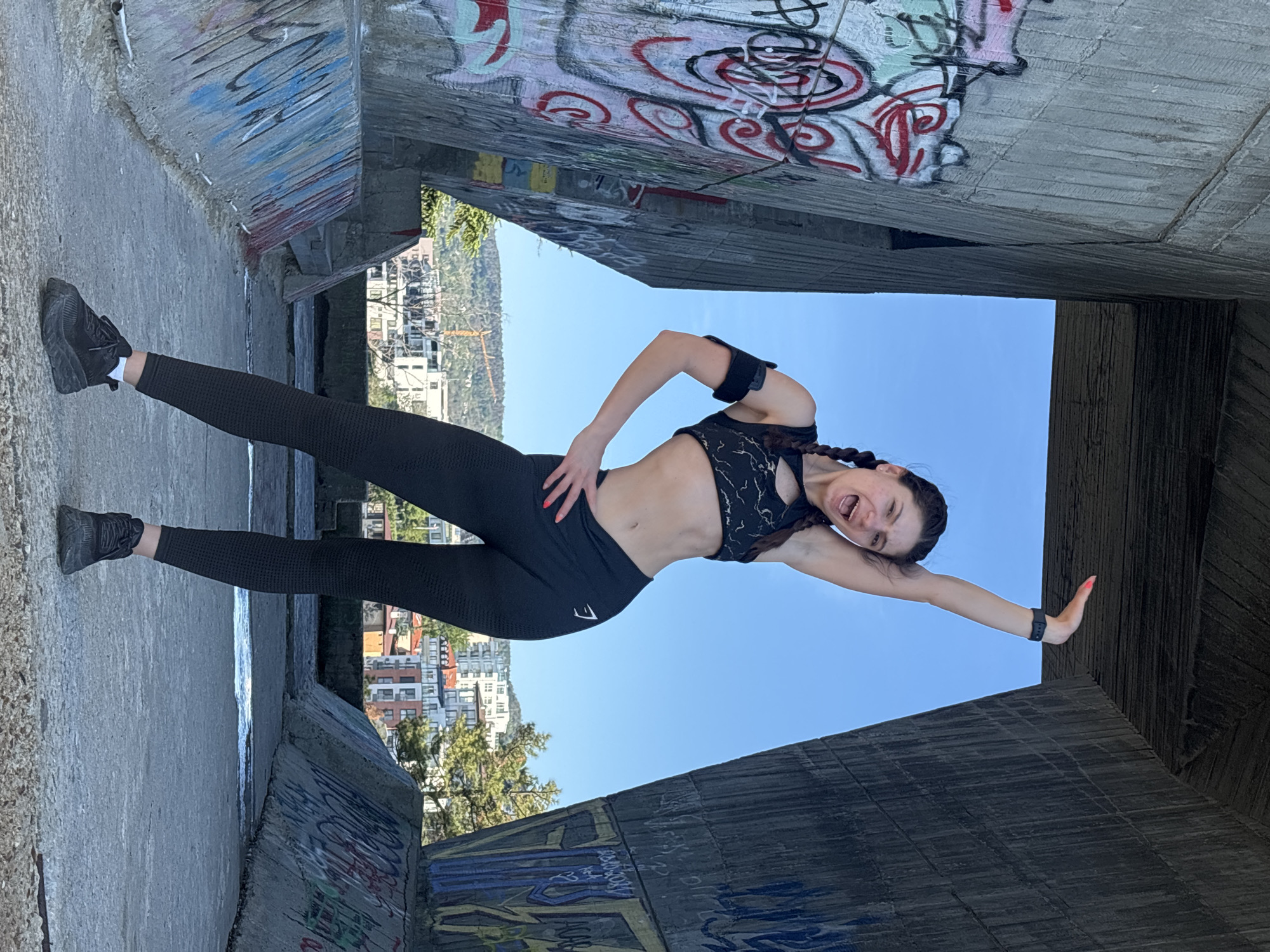 A woman is posing in a split stance with one leg extended to the left and one arm extended to the right, appearing to balance herself between concrete structures. She is wearing black athletic leggings and a black sports bra, and has an armband on her left arm. The background shows a cityscape with buildings and greenery, under a blue sky. The concrete structures are covered in graffiti. The scene is likely a bridge or underpass, judging by the structural elements. The cityscape and architecture suggest the location might be Varna, Bulgaria.