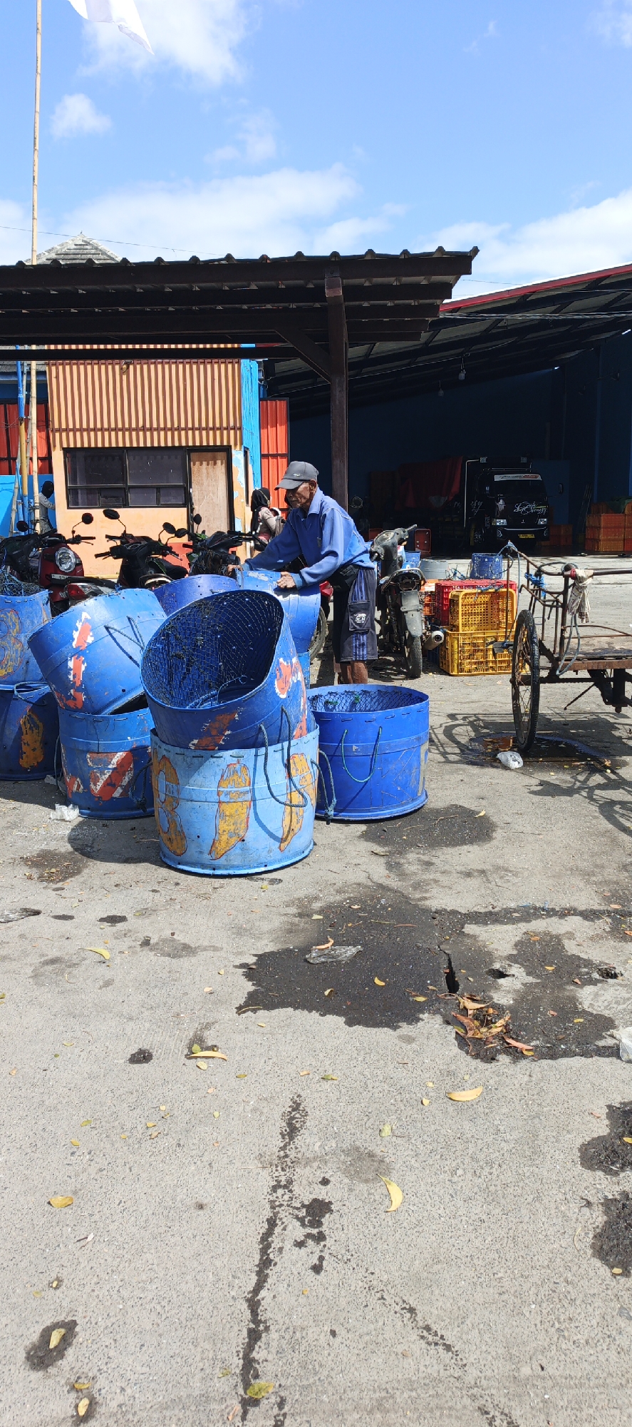 In Puger, Indonesia, a man wearing a blue shirt, black cap, and shorts stands near a pile of large blue cylindrical containers, some of which are decorated with yellow and red images that appear to be stylized bananas or peppers. He is leaning against one of the containers and seems to be working with them. Several motorbikes are parked behind the containers. In the background, there is a building with an orange and brown striped facade, a dark blue warehouse-like structure, and stacked yellow crates. A handcart with two wheels is visible to the right of the scene. The ground is paved and shows several wet, dark patches, possibly from water or oil. A white flag on a bamboo pole is visible in the upper left corner of the image. The sky is blue with scattered white clouds.