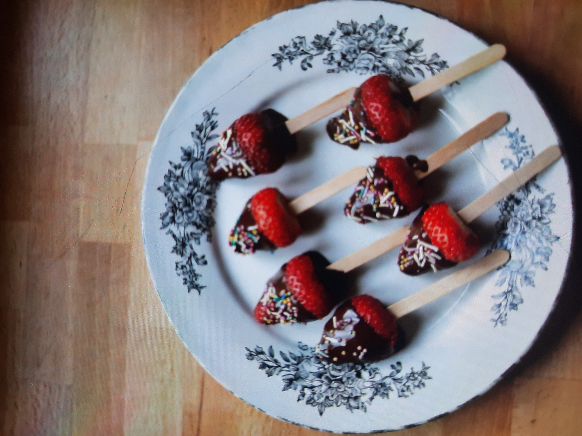An overhead shot shows seven chocolate-dipped strawberries with sprinkles on sticks arranged on a decorative white plate with a floral pattern. The plate is on a wooden surface, likely a table. The strawberries are positioned in a slightly irregular cluster, with most of their red surface visible below a dark brown chocolate coating. Each chocolate-dipped portion is adorned with colorful sprinkles. The plate itself has a dark, ornate floral design around its rim. The lighting suggests an indoor setting. There are no people present in the image, and no specific landmarks or context to place it in Rosenheim, Germany.
