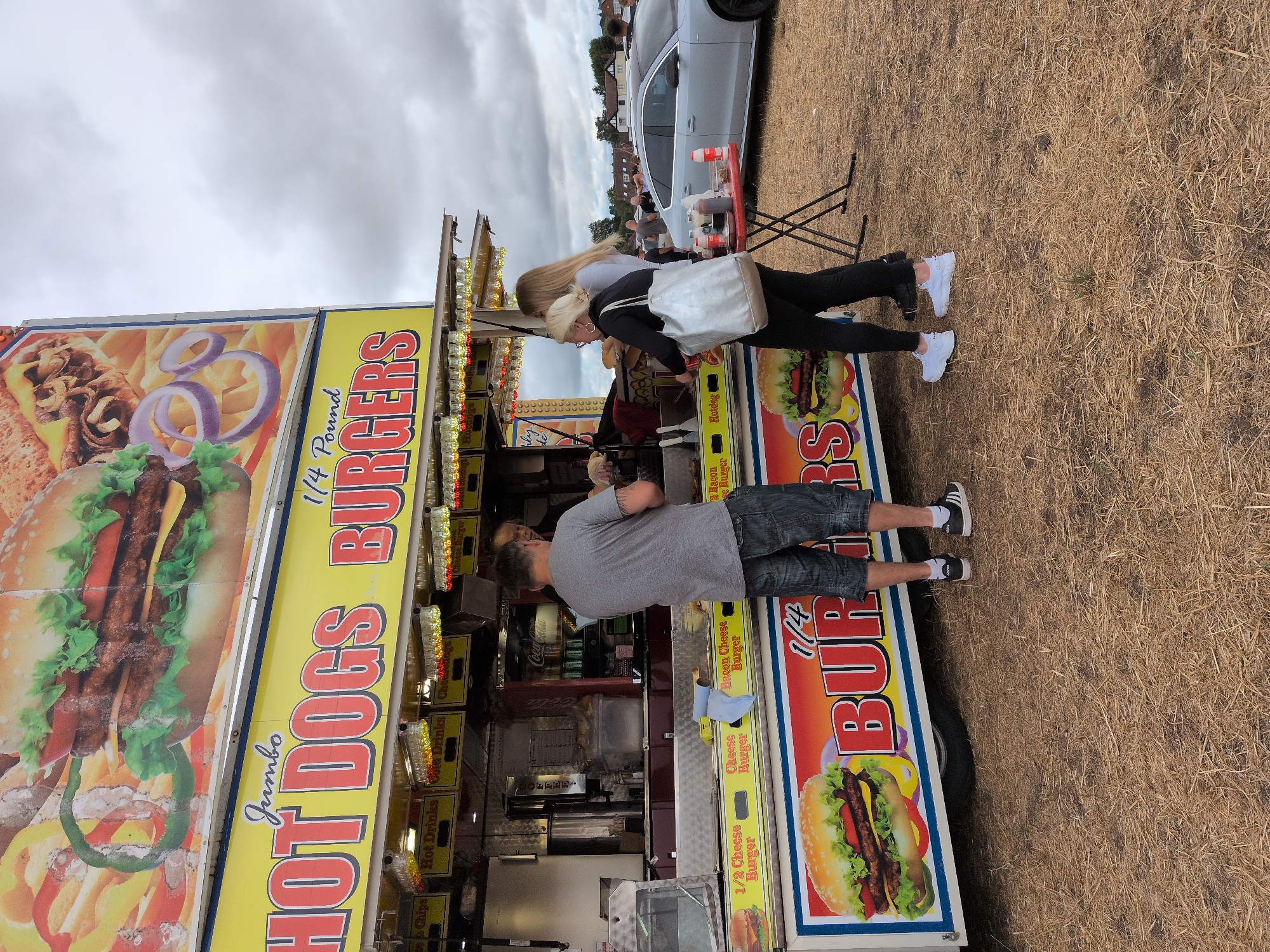The image depicts a food truck selling hot dogs and burgers in South Staffordshire, United Kingdom. A man in a grey t-shirt and denim shorts is ordering food at the counter, while two women stand behind him. A red table with condiments is set up to the right of the food truck. A silver car is parked in the background. The ground is covered in dry grass, suggesting an outdoor event or fair. The sky is overcast, with a few scattered clouds. The food truck features large advertisements for "Jumbo Hot Dogs" and "1/4 Pound Burgers," with images of food items.
