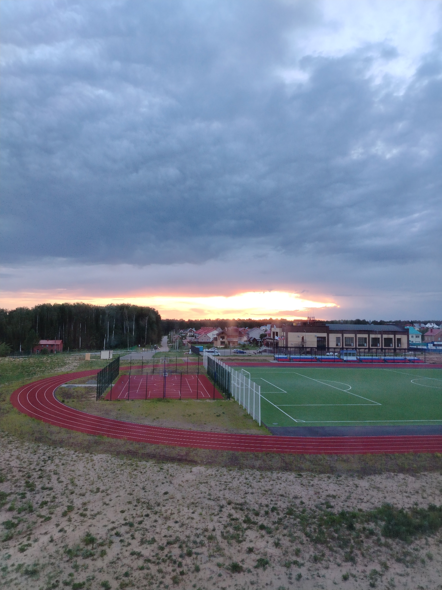 The image depicts an athletic complex in Noviy, Russia, at dusk. The complex includes a red running track, a red basketball court, a green soccer field, and a multi-story building. The sky is overcast with a sunset visible on the horizon. A forest borders the complex on one side, and houses are visible in the background. The foreground shows sandy ground with sparse vegetation. No people are visible.