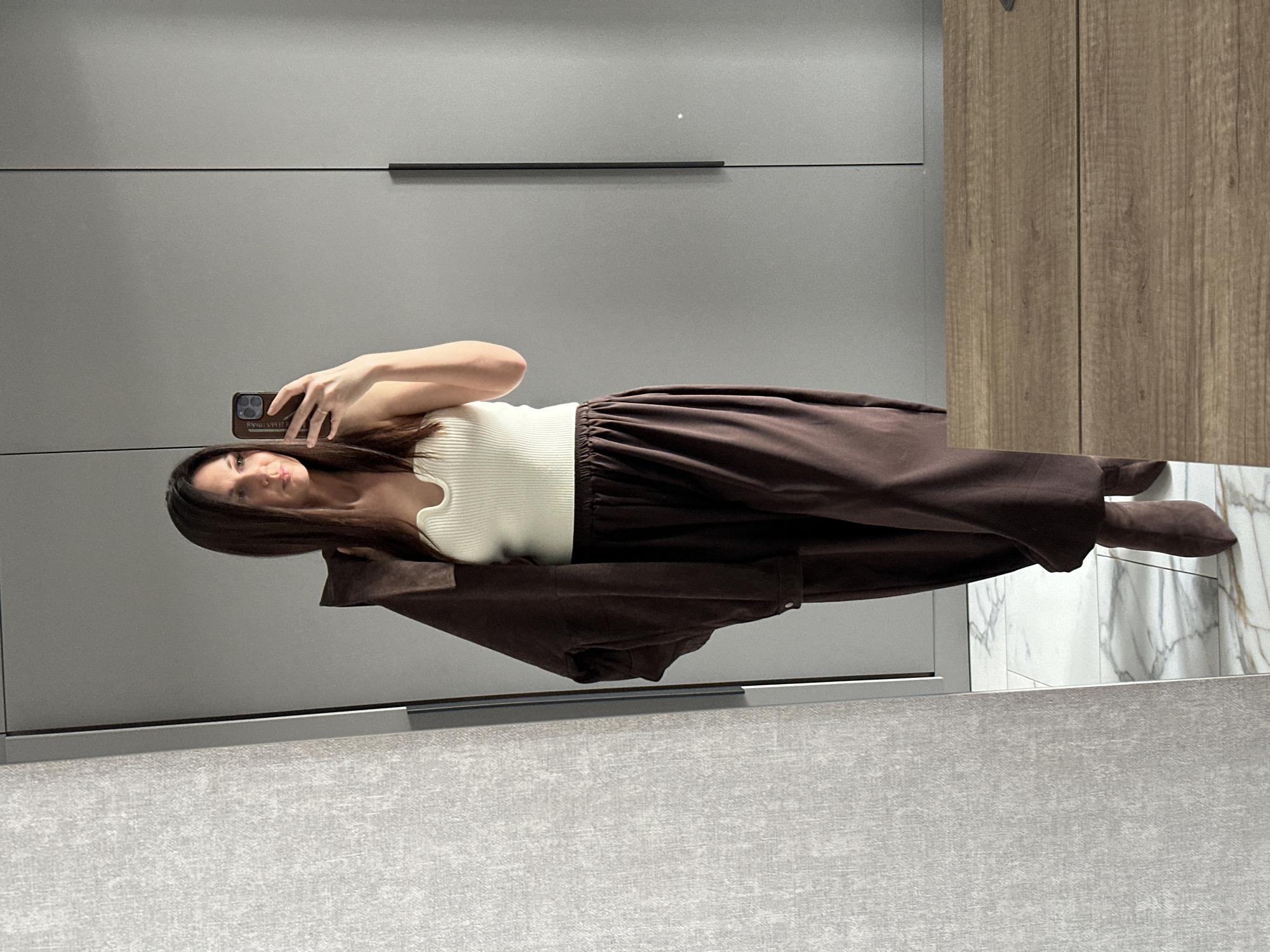 An image taken in Chaikovsky, Russia, depicts a female subject taking a mirror selfie indoors. The female has long, dark brown hair and is dressed in a cream-colored ribbed tank top with a notched neckline, dark brown loose-fitting trousers, and dark brown suede ankle boots. A dark brown jacket, matching her trousers and boots, is draped over her left shoulder. She wears a ring on her left ring finger and holds a smartphone with a dark brown case in her right hand, positioned to capture her reflection in the mirror. Her gaze is directed towards the phone's screen. The immediate environment includes a large mirror on the left, reflecting a textured light grey wall. To the right, grey built-in wardrobes or cabinets with black vertical handles are visible. The floor is covered with white marble-patterned tiles featuring grey veining. In the foreground, a light brown wooden surface, possibly a dresser, is reflected in the mirror.