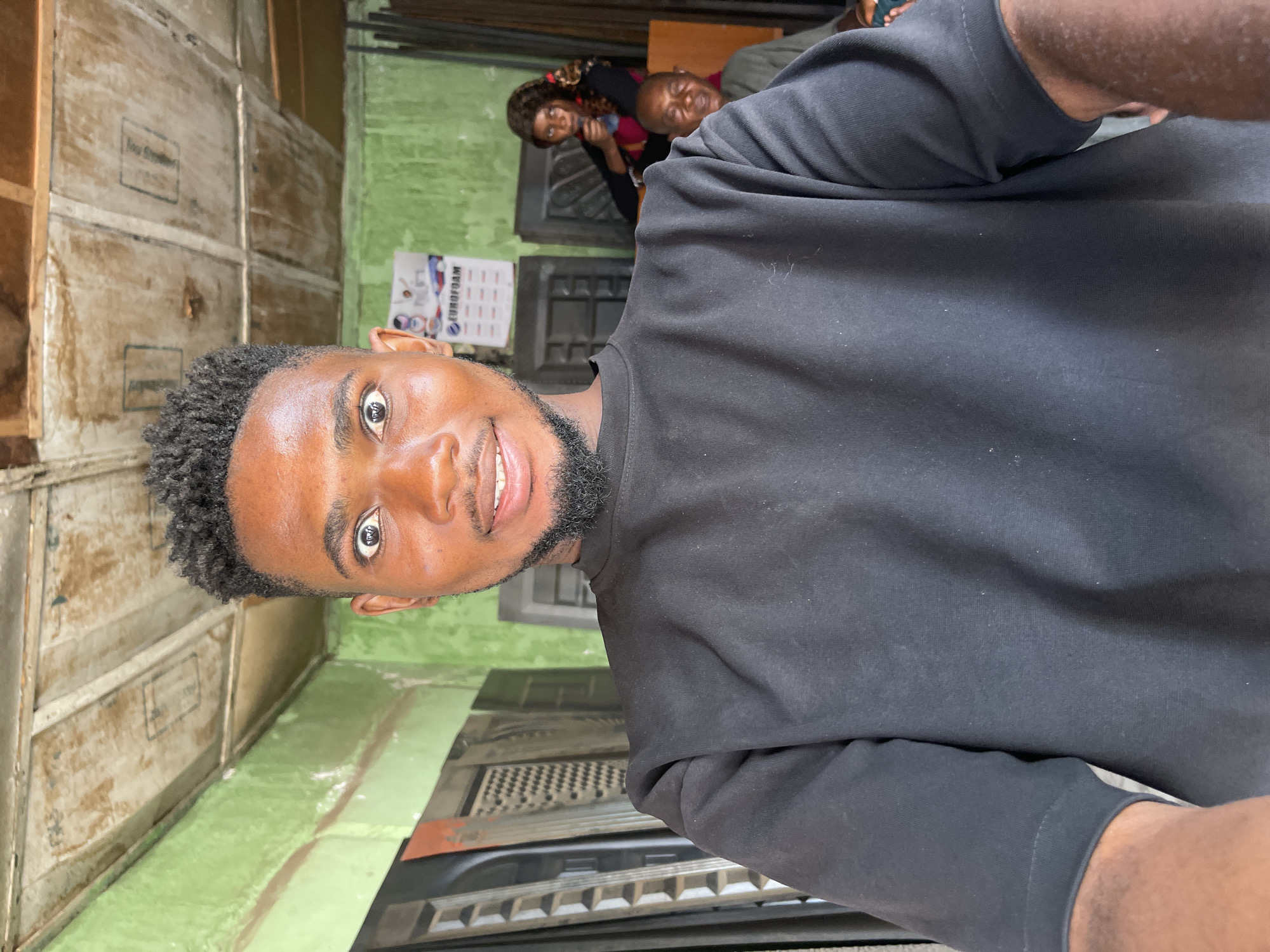 A portrait-style shot captures a young Black man looking directly at the camera with a bright smile. He has short, curly dark hair and a trimmed beard, and is wearing a plain black long-sleeved shirt.
The setting appears to be an informal indoor or semi-enclosed space, consistent with locations found in Zaria, Nigeria. The background is somewhat cluttered and suggests a functional environment. To the left, a wall is covered with multiple worn, light-colored wooden panels or crates, some bearing faint, unreadable text. Adjoining this, a section of the wall is painted a faded light green.
In the mid-ground, behind the main subject, two other Black individuals are visible but out of focus. One is an older man, partially seen, looking in the general direction of the camera. The other is a woman with dark, curly hair, wearing a patterned top, who appears to be holding an object near her face.
On the green wall, a calendar or poster is visible, clearly displaying the brand name "EUROFOAM" along with other small, indistinct text and a grid pattern. In the lower part of the frame, near the main subject's left arm, what looks like the front grille and hood of a vehicle can be discerned, suggesting the space might be a garage, workshop, or a similar commercial area.
The lighting is bright and even, indicating it is daytime. The main subject's visible smile conveys a cheerful and welcoming demeanor.
