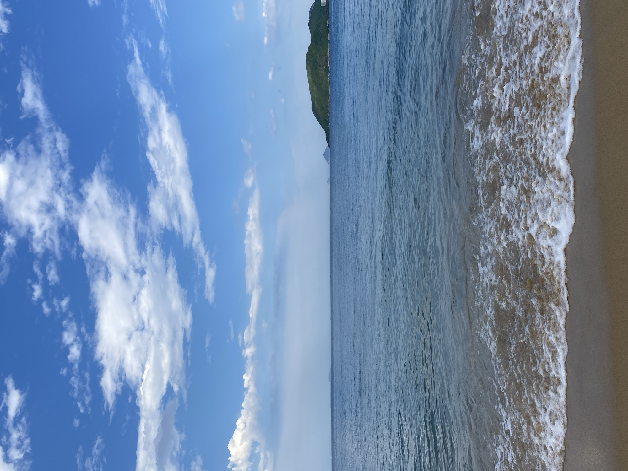 This is a photograph of a beach scene in Nha Trang, Vietnam, taken in daylight under a clear blue sky with scattered white clouds. The image is oriented vertically, with the sky occupying the left half and the sea and beach on the right.
The sky is a vibrant blue, with wispy white clouds, some appearing thin and elongated while others are more voluminous. Towards the horizon, a faint haze can be observed.
The sea is a calm, deep blue, with gentle waves lapping at the shore. The surface of the water displays a rippling texture, reflecting the light of the sun. A line of white foam marks the edge where the waves meet the sand.
The beach is composed of fine, light-colored sand. The foreground shows the wet sand where the waves have receded, transitioning to drier sand further up the beach.
In the background, on the right side of the image, a lush green hill rises steeply from the sea. A few structures, possibly buildings, are visible on the upper slopes of the hill. The horizon is a distinct line separating the sea and the sky.
The scene evokes a sense of tranquility and natural beauty. The overall impression is one of a peaceful day at the coast. There is no visible text in the image.
