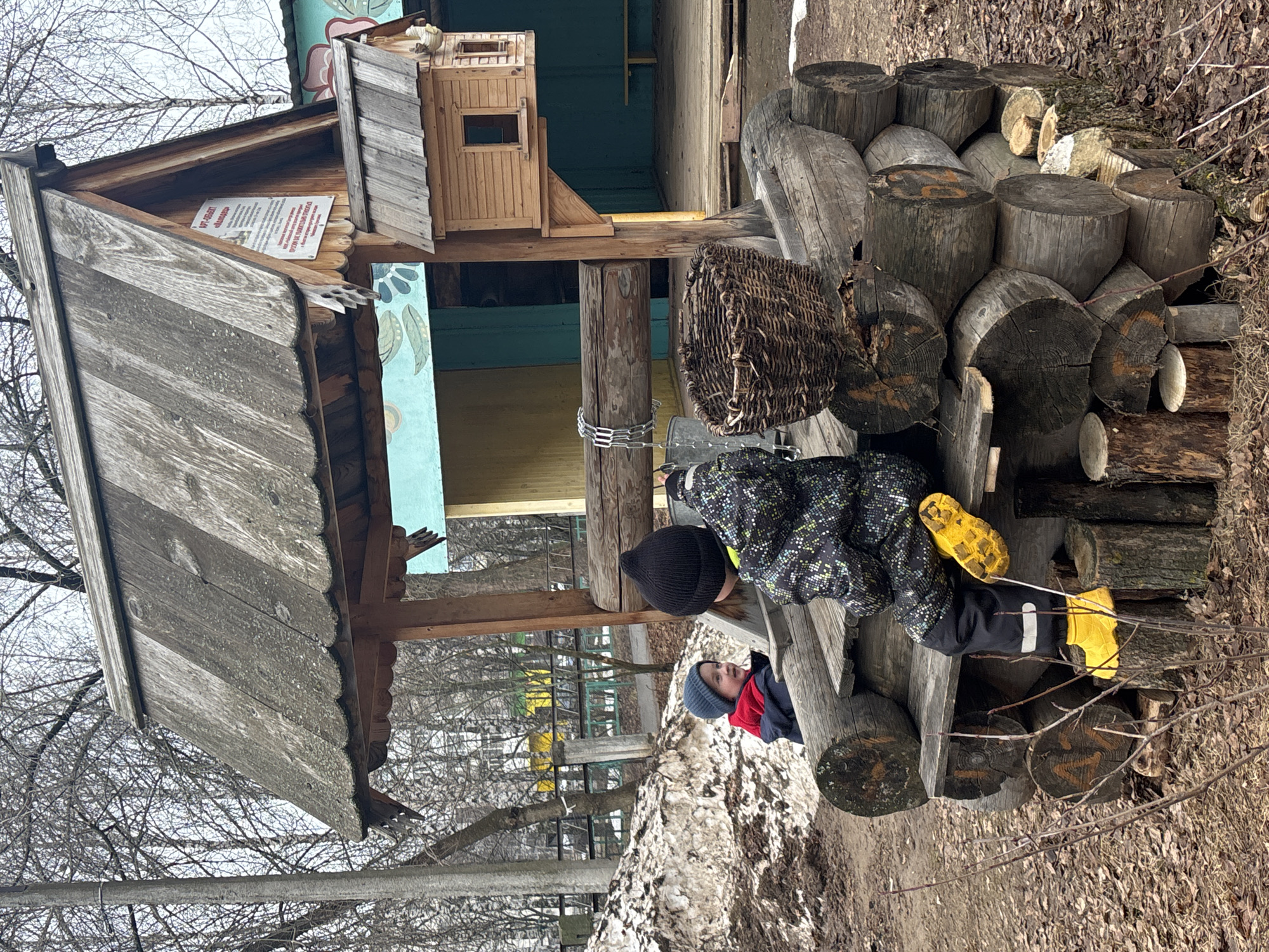 Two children are playing at a playground, with one wearing a black hat and a speckled black and white jacket sitting on a log structure while wearing yellow boots and the other child is visible partially wearing a blue hat and red shirt inside a wooden structure. The playground features logs, a basket, a wooden playhouse with a gray roof and painted walls, along with bare trees in the background, suggesting it might be late winter or early spring. The location is in Chaikovsky, Russia.