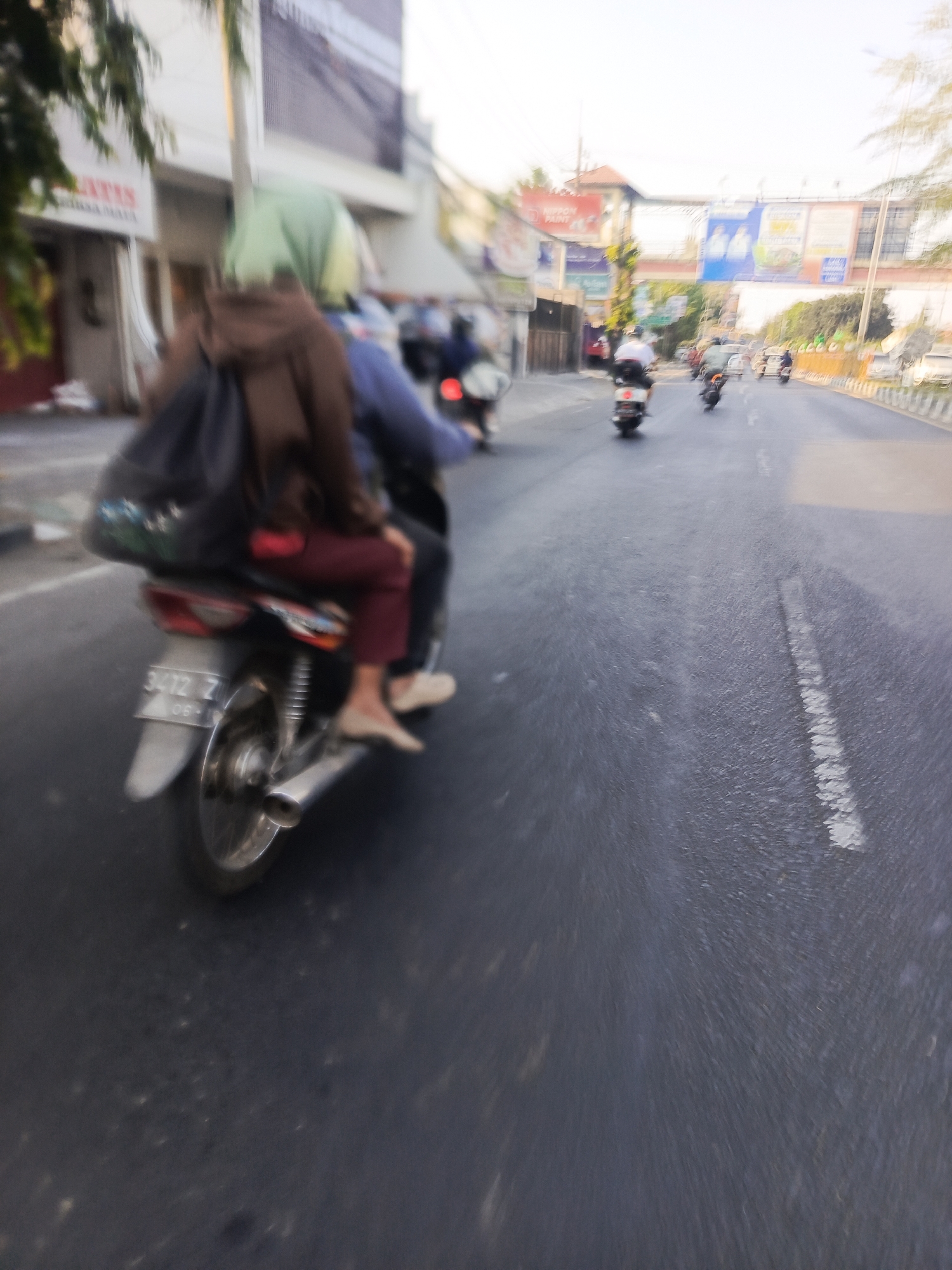The image depicts a motion-blurred scene on a street in Sidoarjo, Indonesia, likely during daylight hours. In the foreground, a motorcycle is seen from the rear, carrying two individuals. The rider, wearing a blue shirt and maroon pants, is partially obscured. The passenger in the back, wearing a light green hijab and a brown jacket, has a dark backpack slung over their shoulder. The motorcycle has a license plate that reads "3412 Z 06-19". The street is paved and appears to be a multi-lane road, with white dashed lines indicating lanes. Other motorcycles are visible further down the road, also in motion. The background shows buildings lining the street, some with signs and awnings. A few trees are also present. The overall impression is a bustling street scene with active traffic.