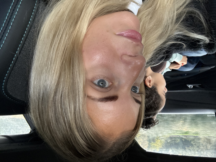 The scene depicts two individuals inside a vehicle. The user-provided context places this scene in Pourrières, France; however, this specific geographic location is not ascertainable from the visual content alone.
In the foreground, a female subject with blonde hair and light blue eyes is partially visible, oriented slightly towards the right of the frame. Her right eyebrow exhibits dark, well-defined hairs, contrasting with her lighter hair. Her lips are light pink. She appears to be seated in a front vehicle seat, with a black seat featuring teal stitching visible to her right. A white garment is partially visible on her person.
In the mid-ground, a younger individual with dark, curly hair pulled back is seated in the rear passenger seat, observed in profile. This individual is looking out the left rear window. They are wearing a light blue or mint-colored long-sleeved top and dark blue trousers. Red nail polish is visible on one of their fingernails as their hands are clasped.
Through the blurred rear window, an exterior environment is visible, characterized by green foliage and a reflective surface, likely water or a wet road, under what appears to be an overcast sky. The vehicle's interior paneling, including a door handle, is dark.