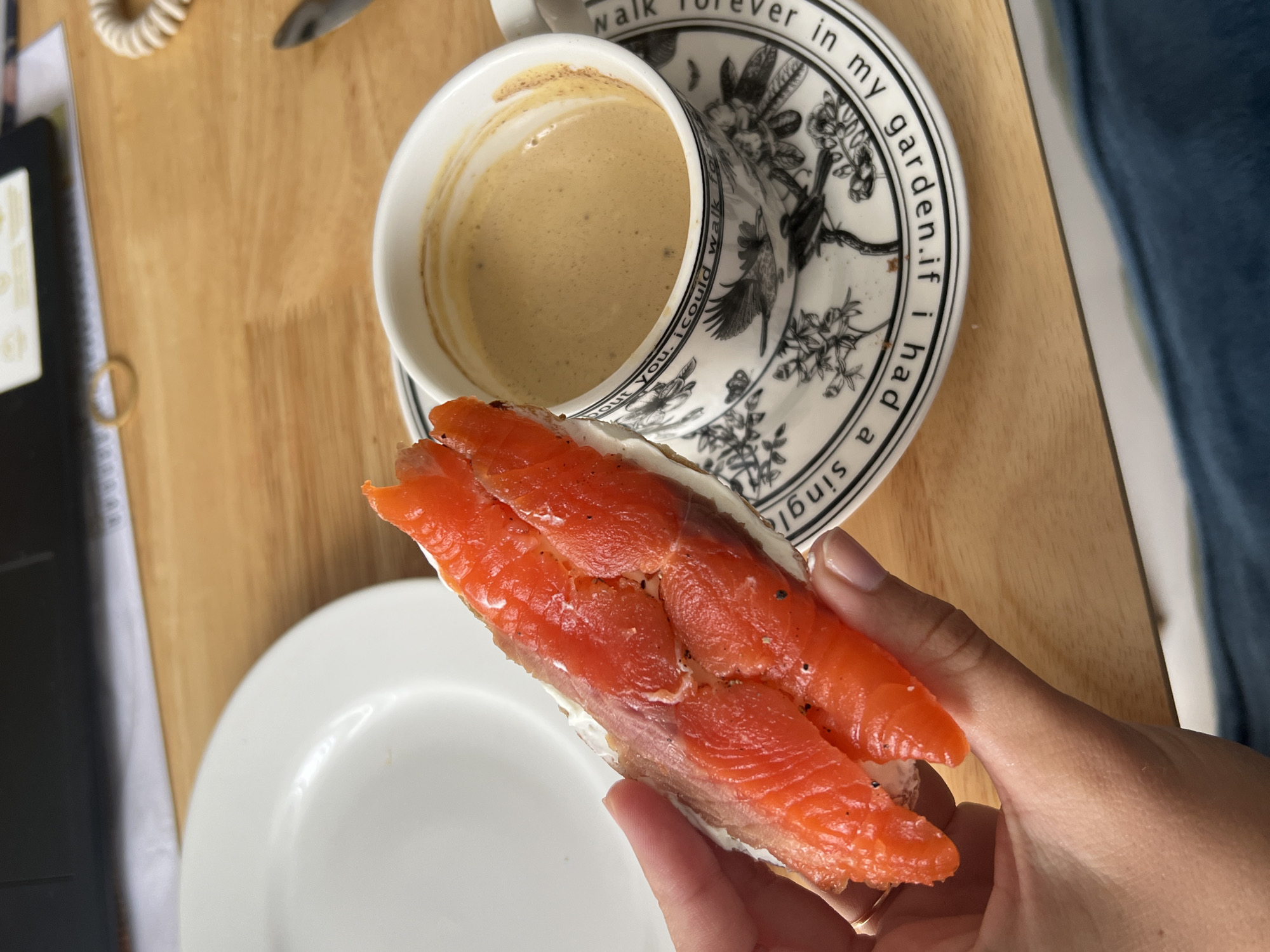 The image shows a person holding a sandwich with salmon and cream cheese. The sandwich is held in the right hand, with the fingertips visible. A white plate is visible in the lower-left corner of the image. A cup of coffee is on a saucer in the upper-center of the image. The saucer has a black and white pattern with a bird and floral design, and text around the rim. The table has a light brown wood grain.