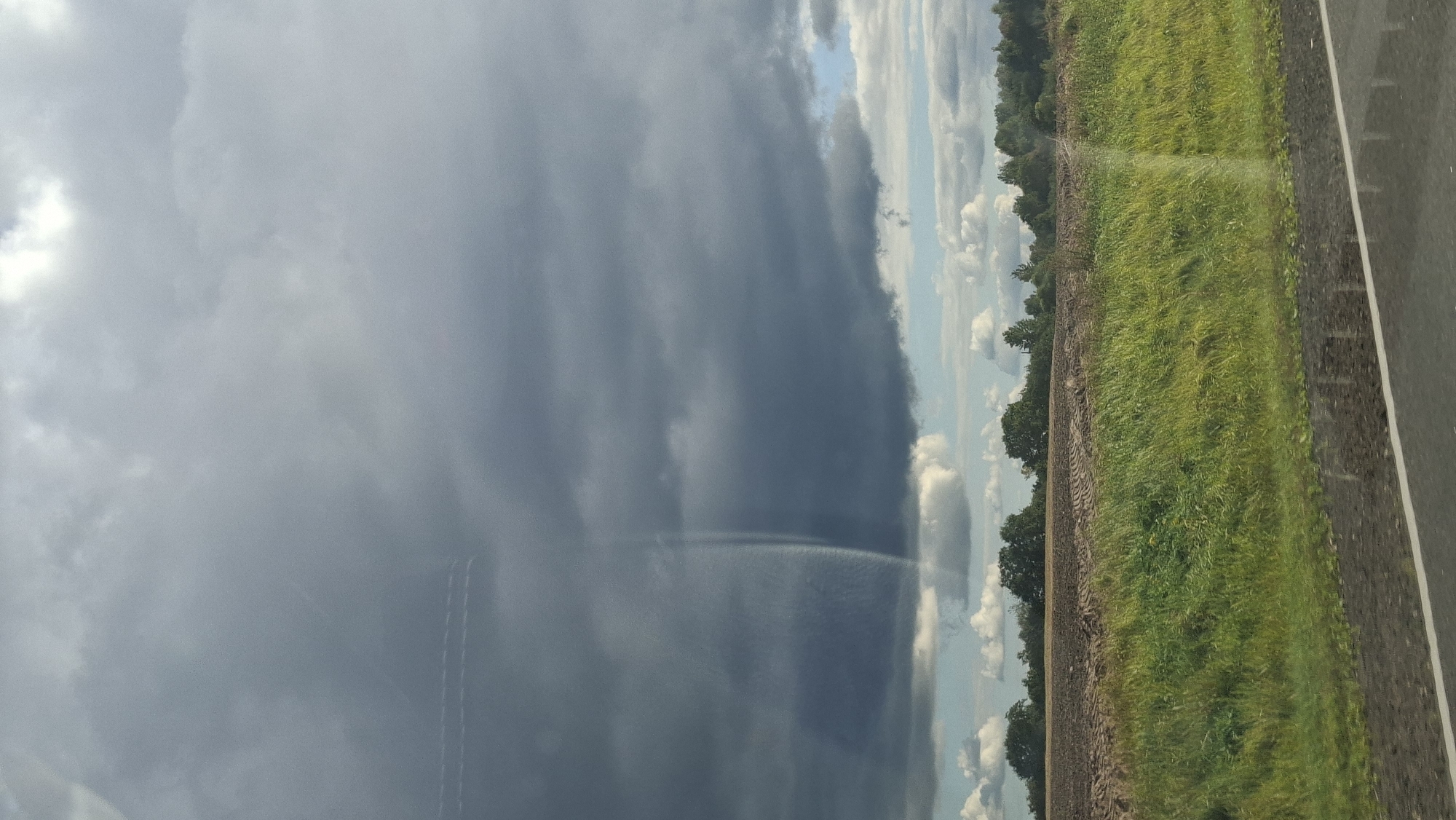 The image shows a rural landscape with a dark, stormy cloud formation in the sky over a field. The foreground features a road with a white line. Beyond the road is a strip of green grass, followed by a brown, plowed field. A line of trees forms the horizon, with more clouds visible in the distance. The overall impression is one of an approaching storm. The location is specified as Верхнякрывінскі сельскі Савет, Belarus. There are no people or animals visible in the image. The scene is static and captured from a fixed perspective, likely from a vehicle due to the road in the foreground. The lighting suggests it is daytime, but the heavy cloud cover creates a dim, dramatic atmosphere.