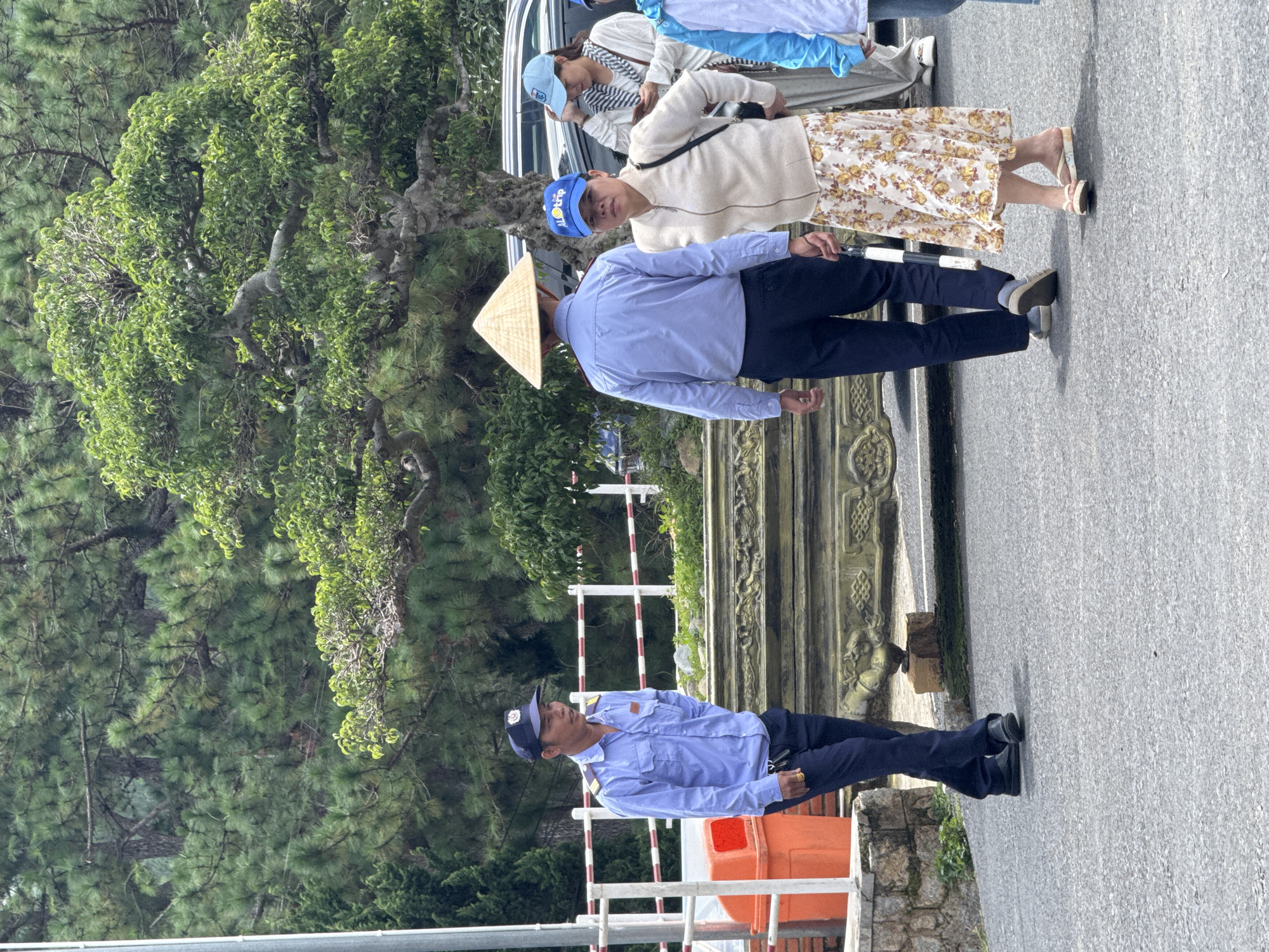 In Da Lat, Vietnam, a man in a blue uniform with a cap stands to the left, looking to his right. He is wearing a light blue collared shirt with epaulets and two chest pockets, and dark blue trousers. He has a ring on his right hand. A large orange trash bin is visible behind him to the left. In the center of the image, a person wearing a conical hat walks away from the viewer, facing right. They are wearing a light blue collared shirt and dark trousers. To the right of this person, a woman in a cream-colored jacket and a floral skirt walks toward the viewer, looking slightly to her left. She is wearing a baseball cap with "ALOTRIP" written on it. Behind her, another woman in a light blue cap and a striped scarf looks at her phone. The background consists of lush green trees and foliage, with a paved road in the foreground.