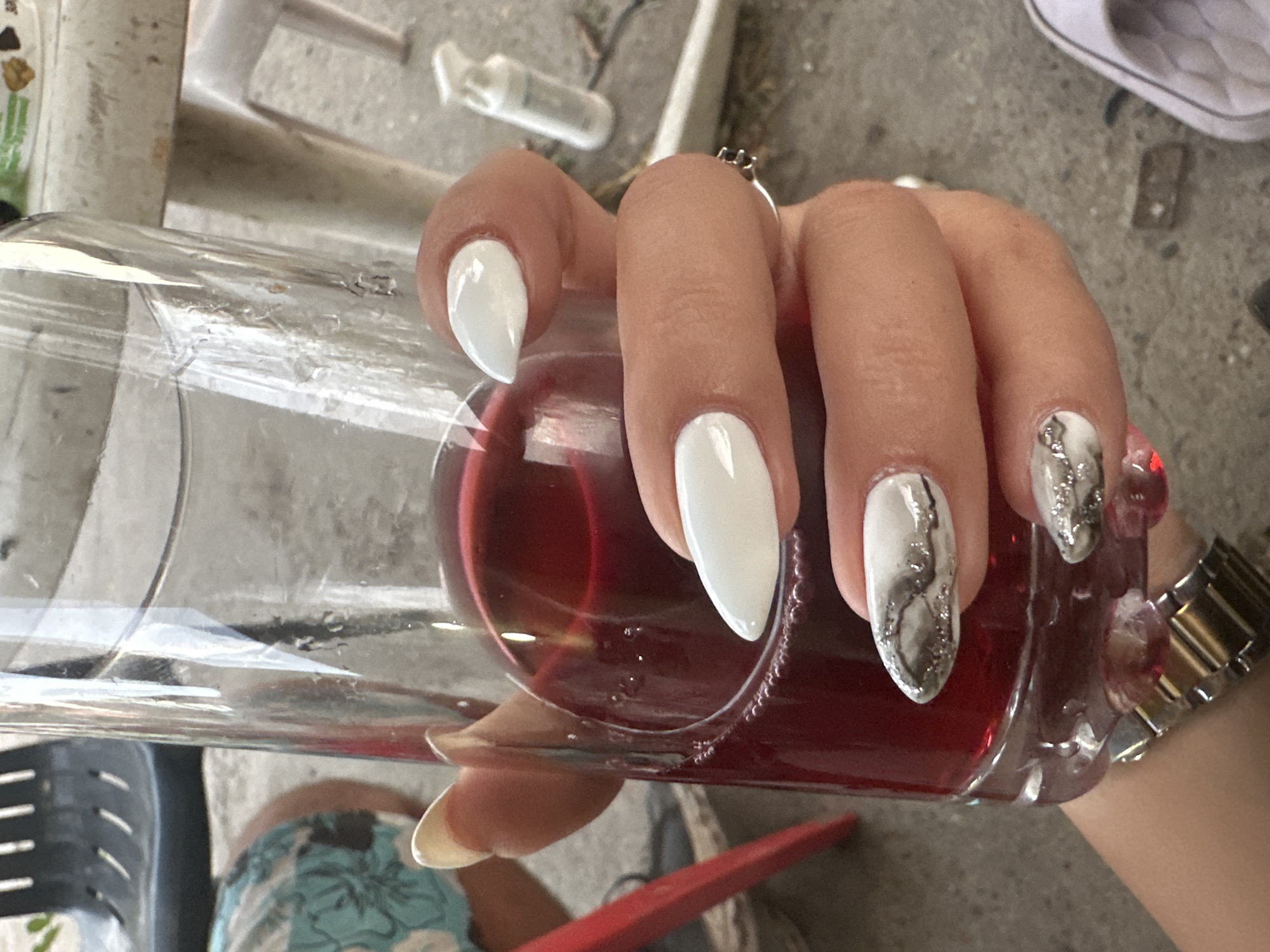 A close-up shot shows a hand holding a glass of red liquid. The hand has long, white acrylic nails, with some of them adorned with a marble-like design of white, grey, and silver glitter. A silver watch is visible on the wrist. The glass contains a red beverage, possibly wine or juice, with some small bubbles clinging to the inside rim. In the background, a black chair with a slatted seat and back is visible, along with parts of a table. The ground appears to be concrete. The scene suggests an outdoor setting, possibly a patio or garden, though no specific urban context or geographical markers are identifiable within the image itself to confirm a location in Galiche, Bulgaria.