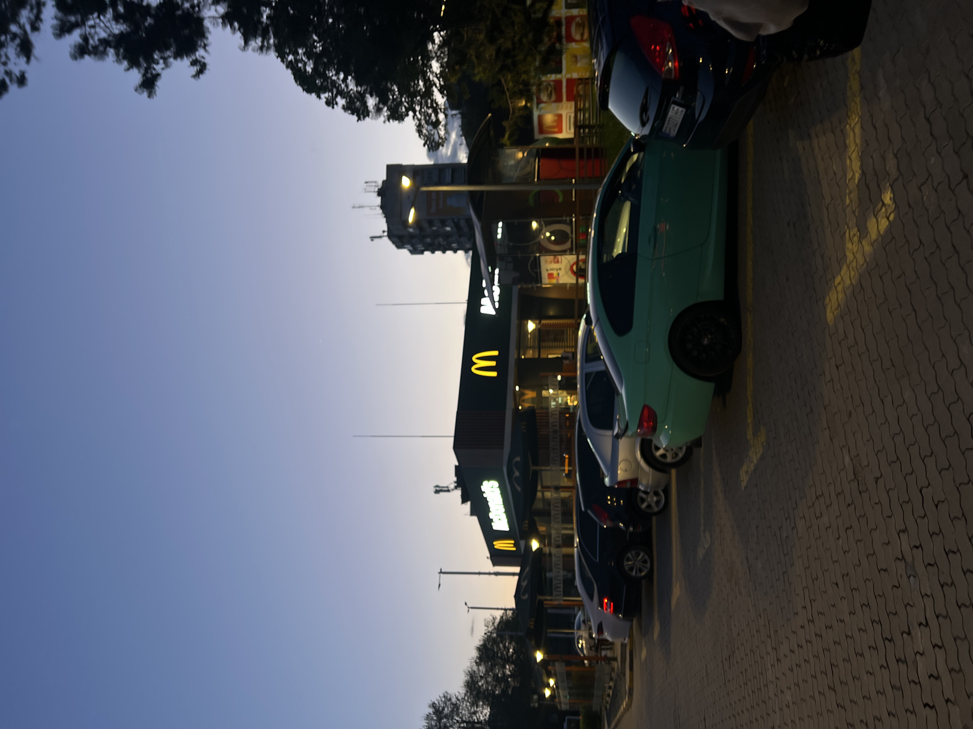 The image captures an evening scene in a parking lot adjacent to a McDonald's restaurant in Plovdiv, Bulgaria. The sky, a gradient of deep blue fading to lighter hues, indicates the time of day is dusk or early evening, with artificial lights beginning to illuminate the surroundings.
In the foreground and to the right, several cars are parked in rows on a paved surface featuring yellow parking lines. A prominent teal or light green car is visible, alongside darker vehicles including black, dark blue, and a silver/white car further down the lot.
The central subject is a brightly lit McDonald's restaurant building, identifiable by its iconic golden "M" arches and "McDonald's" text illuminated in yellow. The building's architecture suggests a modern, commercial design. Beyond the McDonald's, a taller multi-story building, possibly residential or commercial, stands against the darkening sky.
Streetlights are visible, casting light onto the parking area and the buildings. Trees and foliage are present on the left side of the frame and around the buildings, with their silhouettes standing out against the twilight sky. No people are visible in the image, contributing to a quiet, serene atmosphere. The pavement consists of interlocking grey pavers.