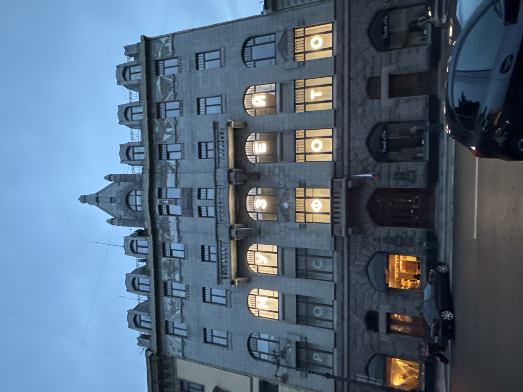 The image captures a historical building in Saint Petersburg, Russia, with letters illuminated in its windows, forming words. The architecture of the building is stately, characterized by stone facade, arched windows, and decorative elements typical of pre-revolutionary St. Petersburg architecture. Several windows on the upper floors display illuminated Cyrillic letters. On one level, the letters "М", "У", "З", "Е", and "Й" are visible, suggesting the word "МУЗЕЙ" (Museum). Below this, on another floor, the letters "Р", "О", "С", "С", "И", and "Я" are illuminated, forming "РОССИЯ" (Russia). The word "ФОТО" (Photo) is also visible in another window. Streetlights are present in front of the building, and cars are parked on the street. The sky is overcast and appears to be twilight. The overall scene suggests a cultural or historical institution.
