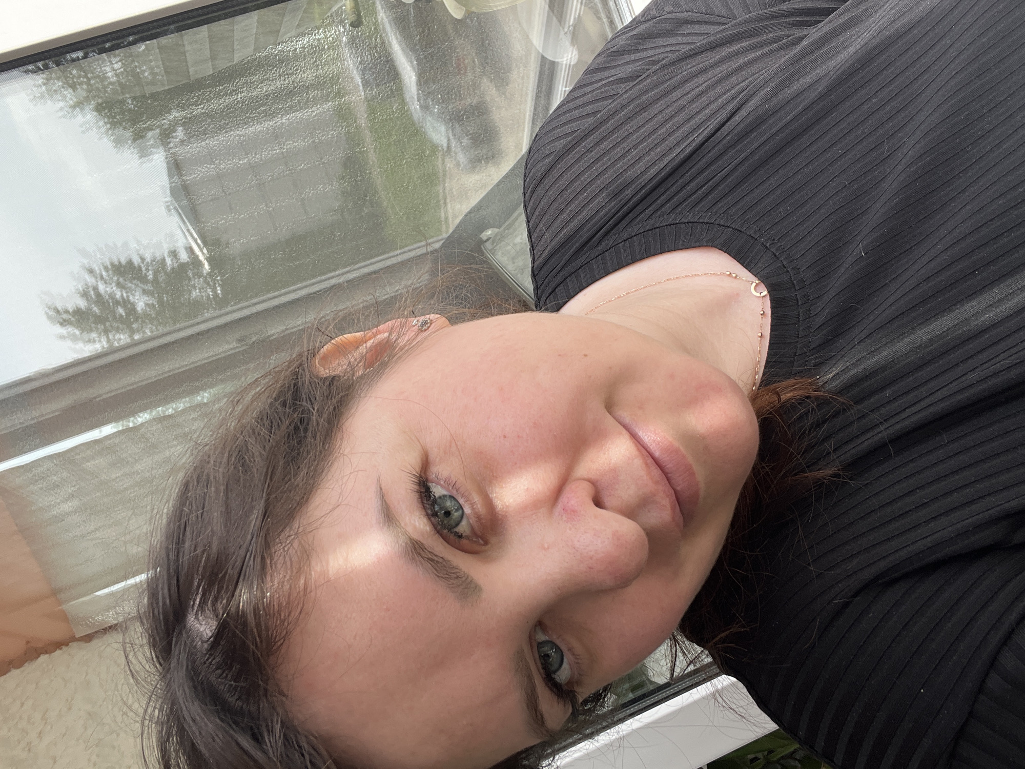 A medium-shot captures a woman with fair skin, light green eyes, and dark brown hair, angled slightly towards the viewer. She wears a black, ribbed shirt and a delicate gold necklace. Her expression is softly smiling. The background appears to be a window, with an indistinct reflection of a tree visible through the glass. The surroundings suggest an interior setting. No specific details connect the image to Noviy, Russia.