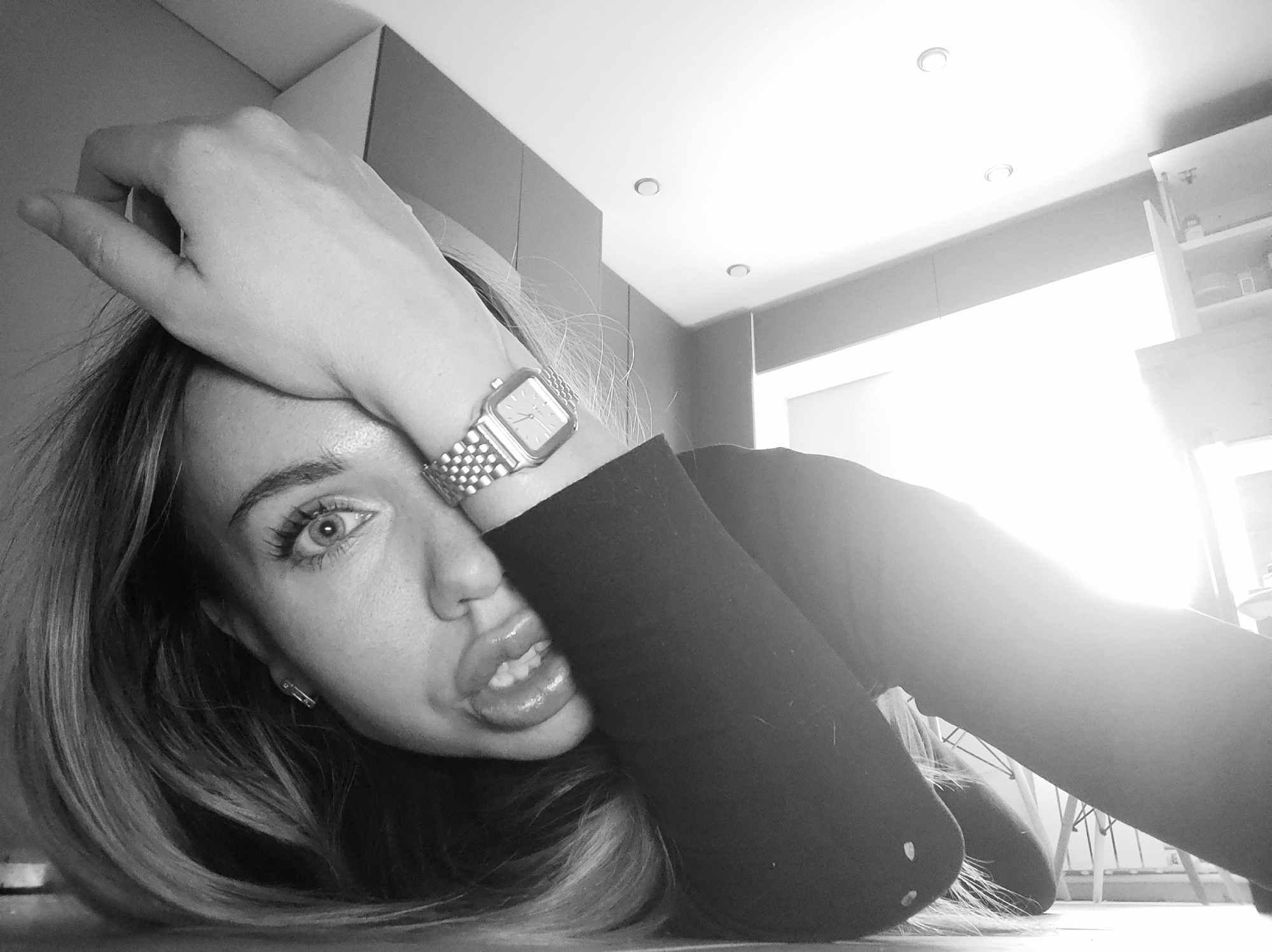 A close-up, grayscale photograph shows a woman lying on a flat surface indoors. She rests her head on the surface with her right hand on her forehead, displaying a silver wristwatch. Her eyes are open, and her lips are slightly parted. She is wearing a dark, long-sleeved shirt. The background includes a white ceiling with recessed lights and gray cabinets. A cabinet is partially open, revealing items inside. The environment and objects do not definitively indicate the city of Votkinsk, Russia.