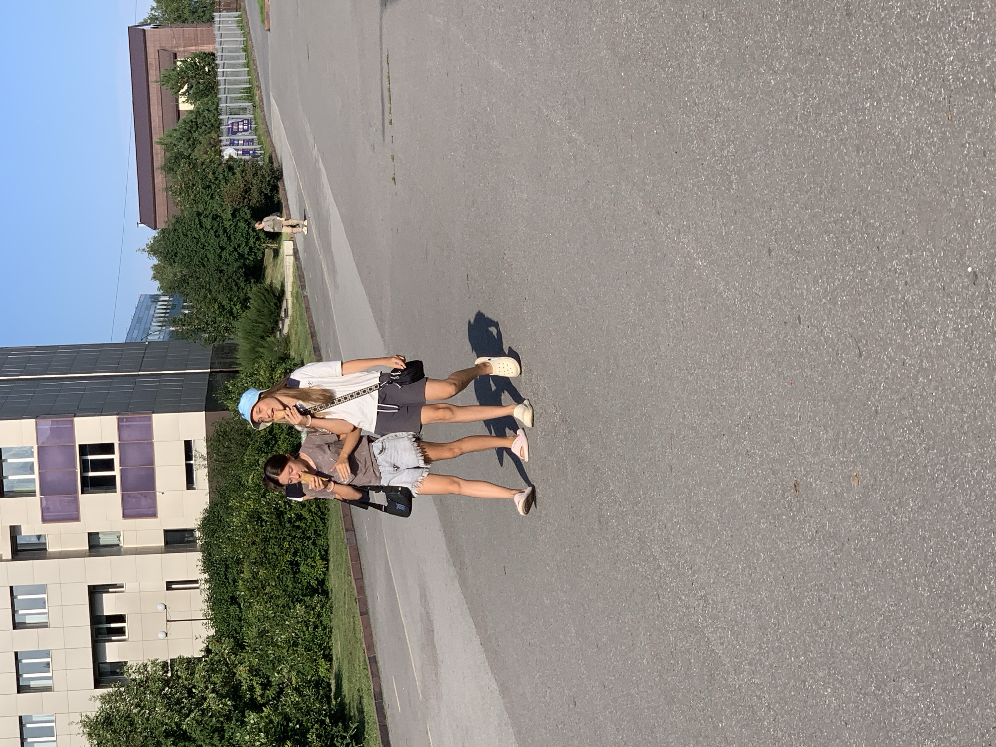 In Prokopyevsk, Russia, two young females are walking down a paved road, each eating an ice cream cone. The female on the left is wearing a gray t-shirt and denim shorts, while the one on the right is wearing a white t-shirt and dark shorts, with a blue cap. Both are carrying small black bags. In the background, a modern, multi-story building with a light-colored facade stands behind a line of green bushes. Further in the distance, another building with a brown roof is visible, along with trees and a fence. The sky is clear and blue.