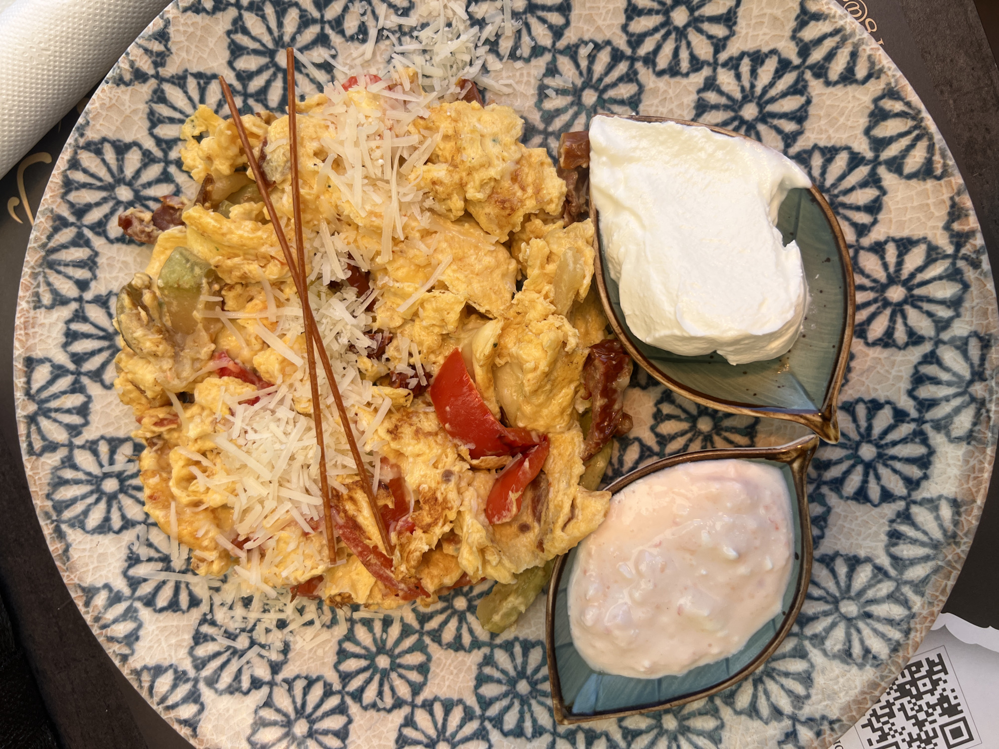 The image shows a plate of food, likely a breakfast dish, on a patterned plate. The main component appears to be scrambled eggs with chopped vegetables such as red peppers and possibly pickles. The eggs are topped with shredded cheese and two decorative brown sticks. Two leaf-shaped dishes on the right side of the plate contain white creamy substances, one possibly sour cream and the other a pinkish-white sauce. The plate sits on a dark surface, possibly a table. A rolled napkin is partially visible in the upper left corner, and a partial receipt with a QR code is visible in the lower right corner. The image was taken in Podgorica, Montenegro.