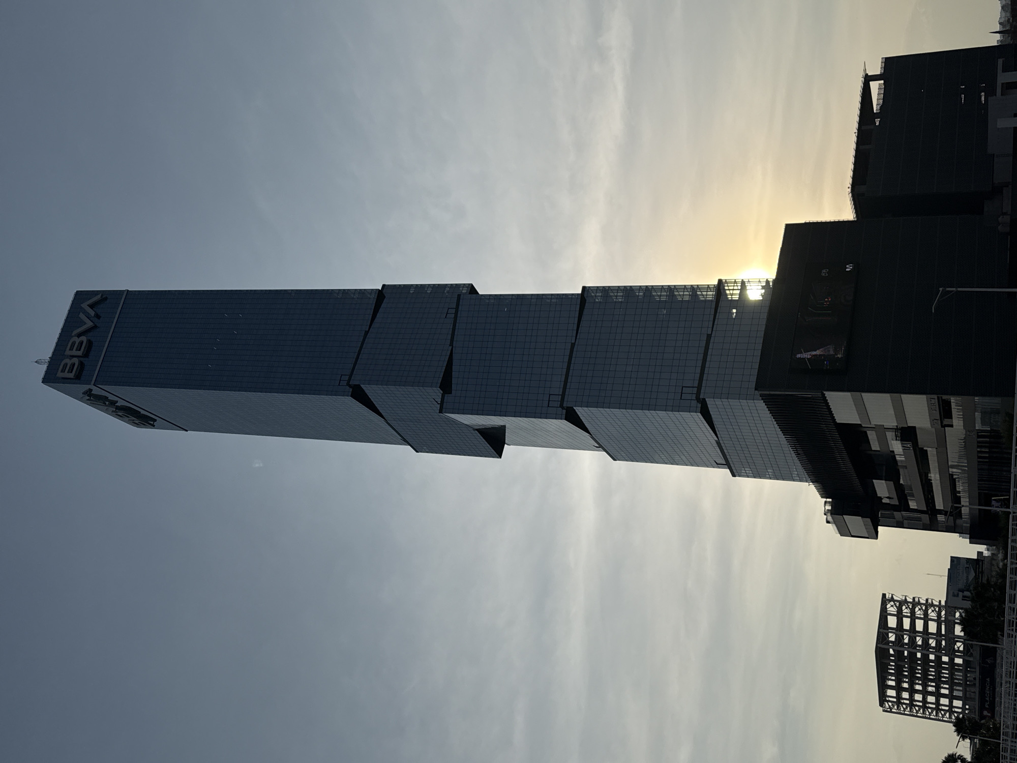 The BBVA tower in Puebla, Mexico, stands tall against a sunlit, cloudy sky. The tower's design features a series of stacked, slightly offset rectangular sections. To the right are other buildings, including one under construction and another with a prominent billboard. The buildings are silhouetted against the sky, obscuring finer details.
