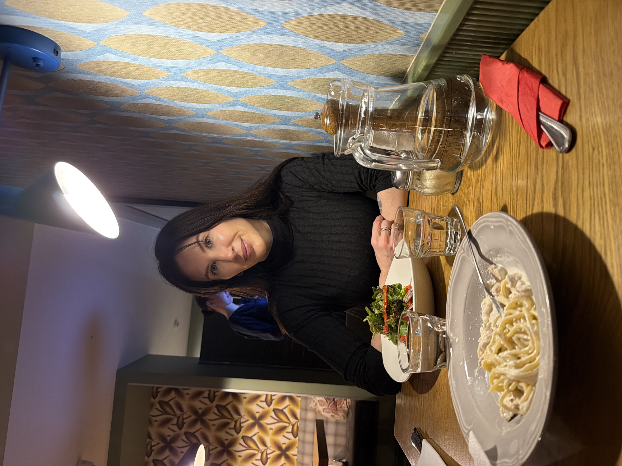 A well-lit interior dining scene, potentially a restaurant, features a woman seated at a wooden table, with pasta and a salad. A glass jug and a red napkin with silverware are also on the table. She is wearing a dark long-sleeved shirt. The location is possibly Perm, Russia.