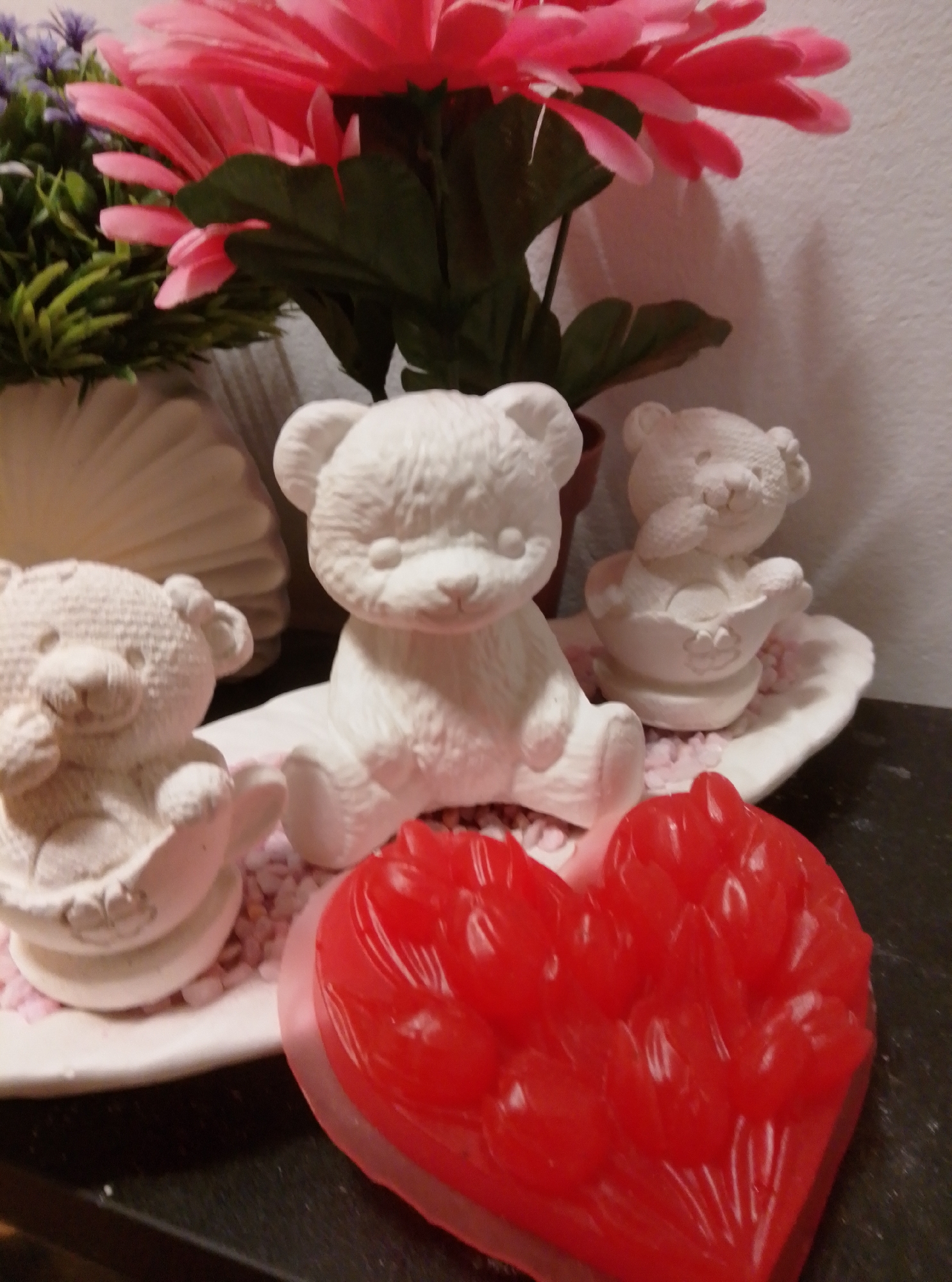 The image shows a still life arrangement on a dark, textured surface. In the foreground, a translucent, red, heart-shaped object with a textured surface is prominently displayed. Behind it, on a white, wavy platter, are three unpainted ceramic teddy bear figures. The largest bear sits in the center, with two smaller bears positioned to its left and right. The bears are surrounded by small, pink, pebble-like objects. In the background, a decorative vase holds artificial pink and purple flowers with green foliage. The scene is illuminated by soft lighting, casting subtle shadows. There is no indication of the city of Wedel, Germany, or any specific location within it.