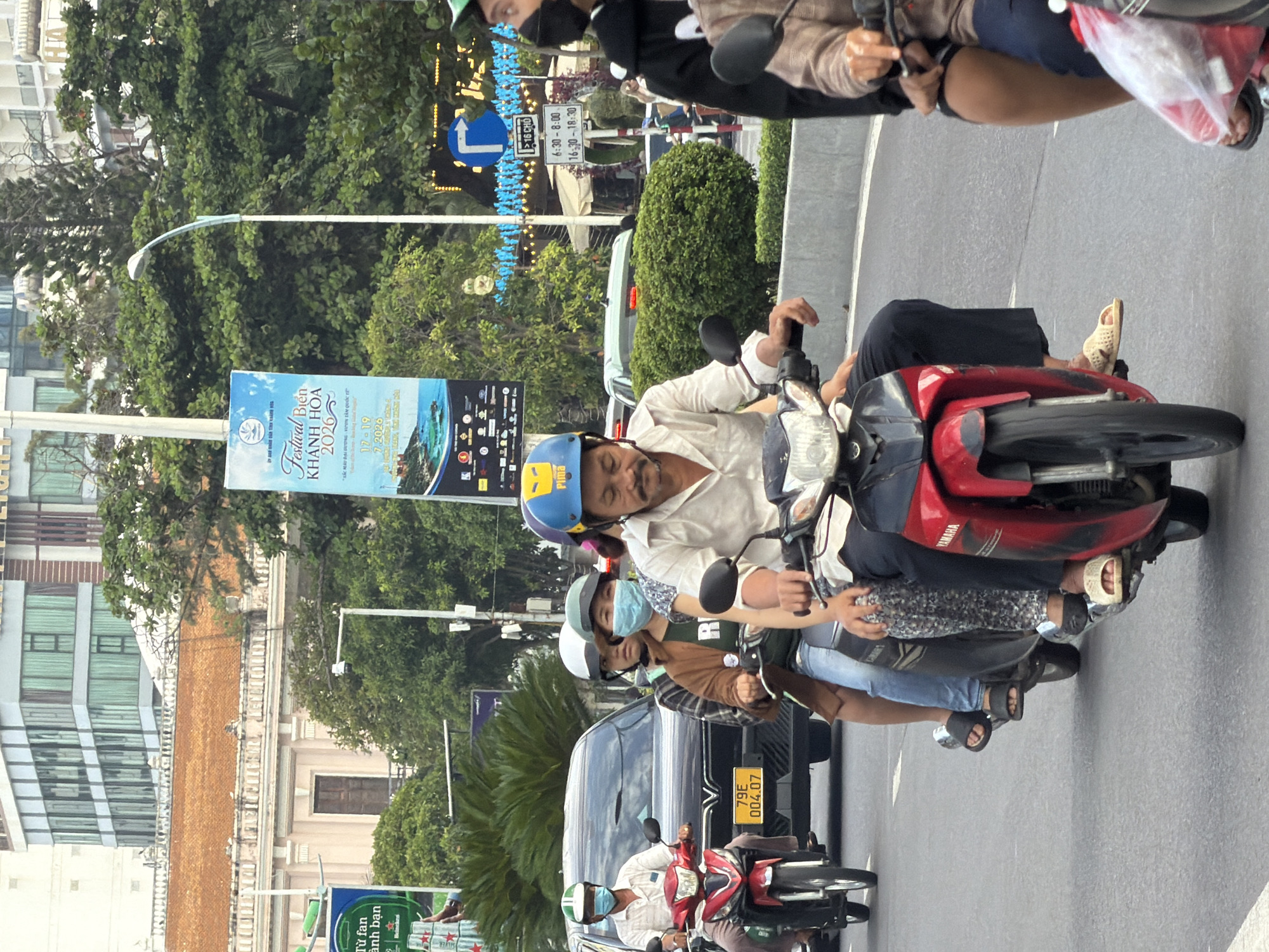 On a city street in Nha Trang, Vietnam, several people ride on motorbikes. The man in the foreground is wearing a light-colored shirt, a blue helmet, and a mustache, and is operating a red and black Yamaha motorbike. Behind him, a woman with a blue face mask is riding on the same motorbike, wearing a brown jacket and jeans. A third person, wearing a patterned headscarf and dark clothing, is also on the motorbike. In the background, a black car is visible. To the left, another motorbike carries two people. The street is paved, and there are trees and buildings in the background, including a banner advertising "Festival Bien Khanh Hoa 2026." The overall scene depicts typical urban traffic in Vietnam.