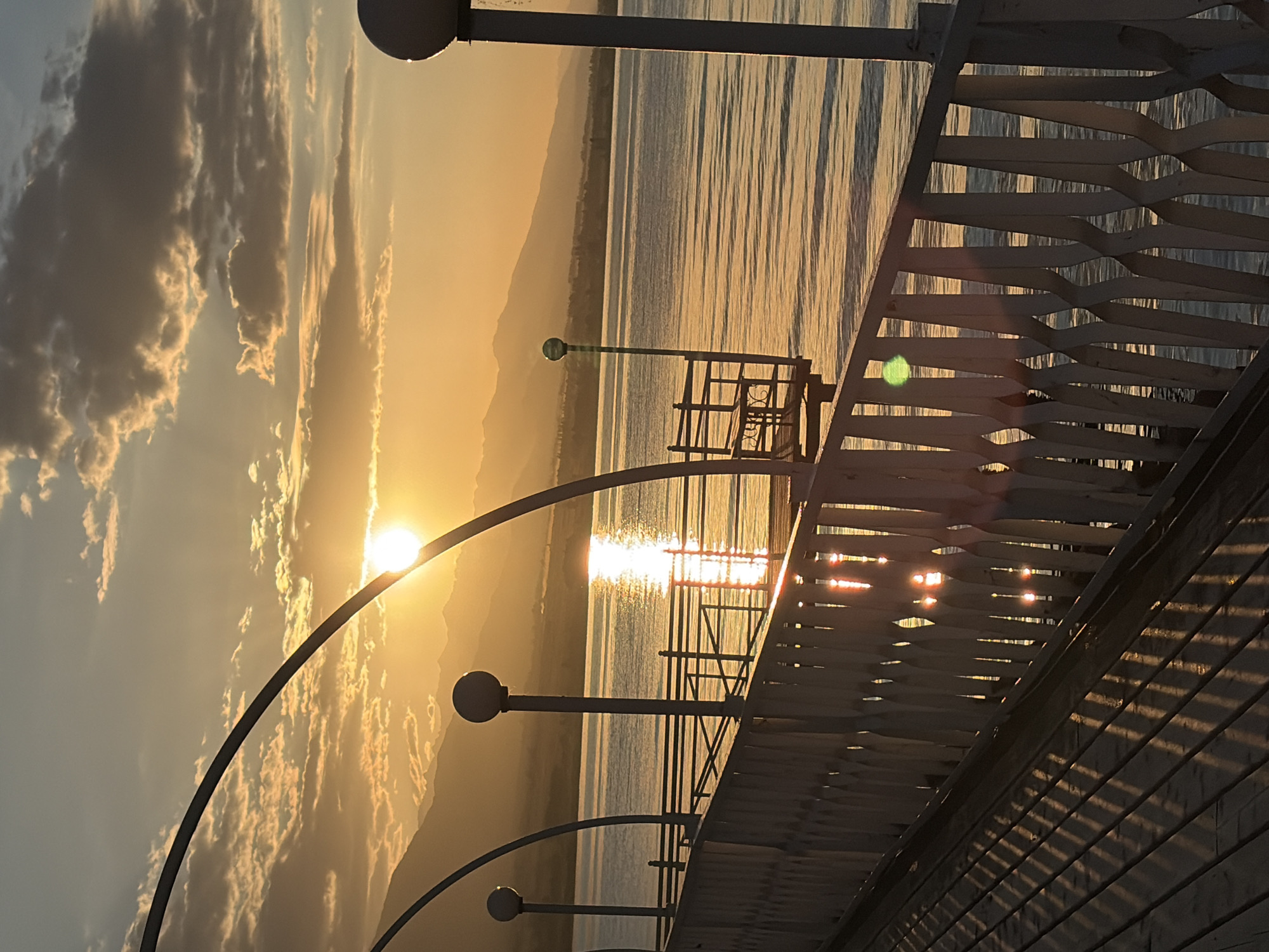 A sunset over a body of water is depicted, with a pier in the foreground. The sun is low on the horizon, casting a warm, golden light across the scene. The sky is filled with clouds, some of which are illuminated by the setting sun. In the distance, mountains can be seen. The pier is made of wood and has a white railing. There are streetlights on the pier. The water is calm and reflects the colors of the sky.