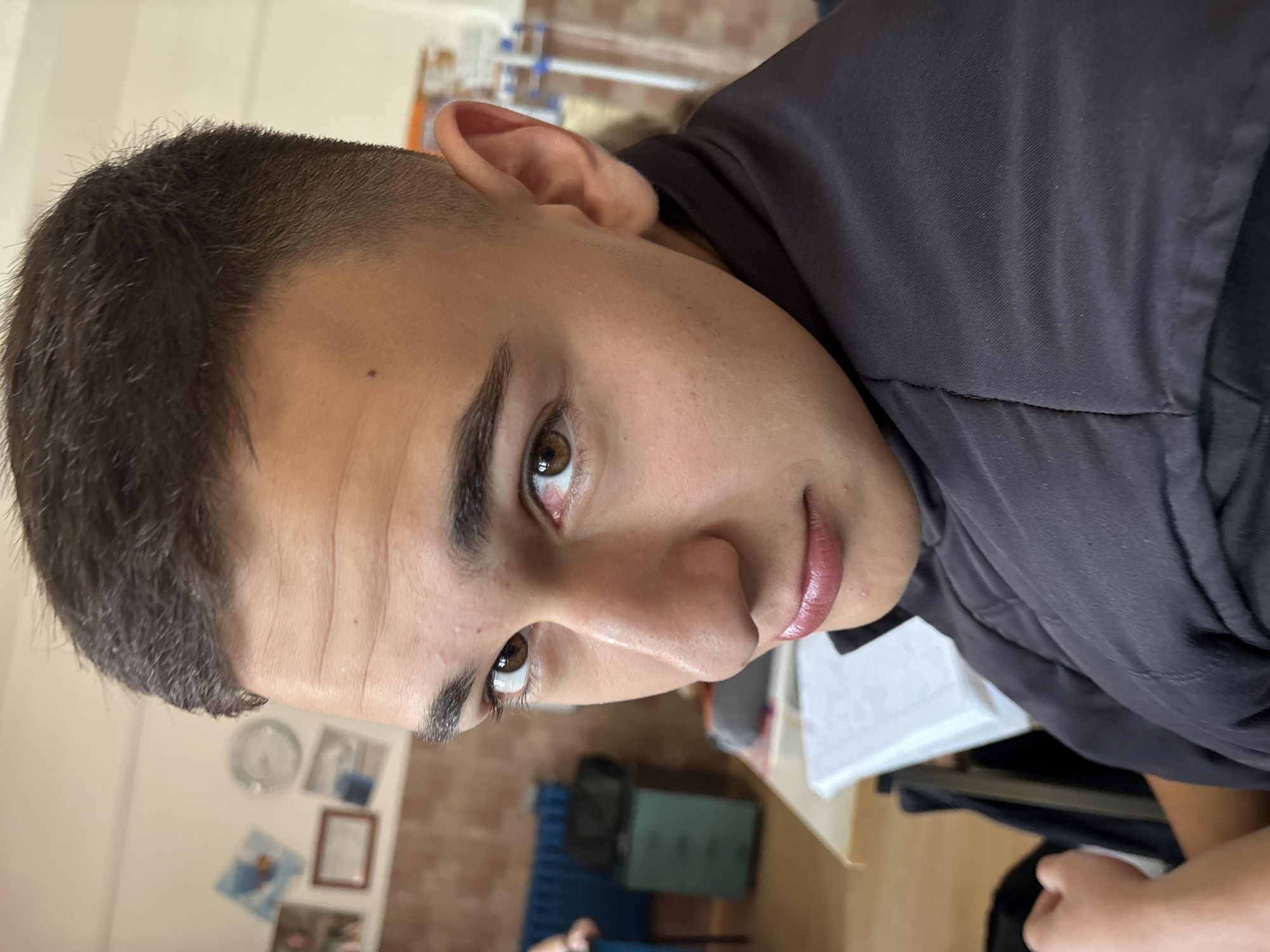 A young man with brown eyes and short dark hair is photographed indoors. He is wearing a dark-colored shirt or jacket. The background includes a wall with framed pictures, a clock, and a blue object, possibly a cabinet or storage unit. There are papers on a surface nearby. The location is likely an interior space in Panagyurishte, Bulgaria.