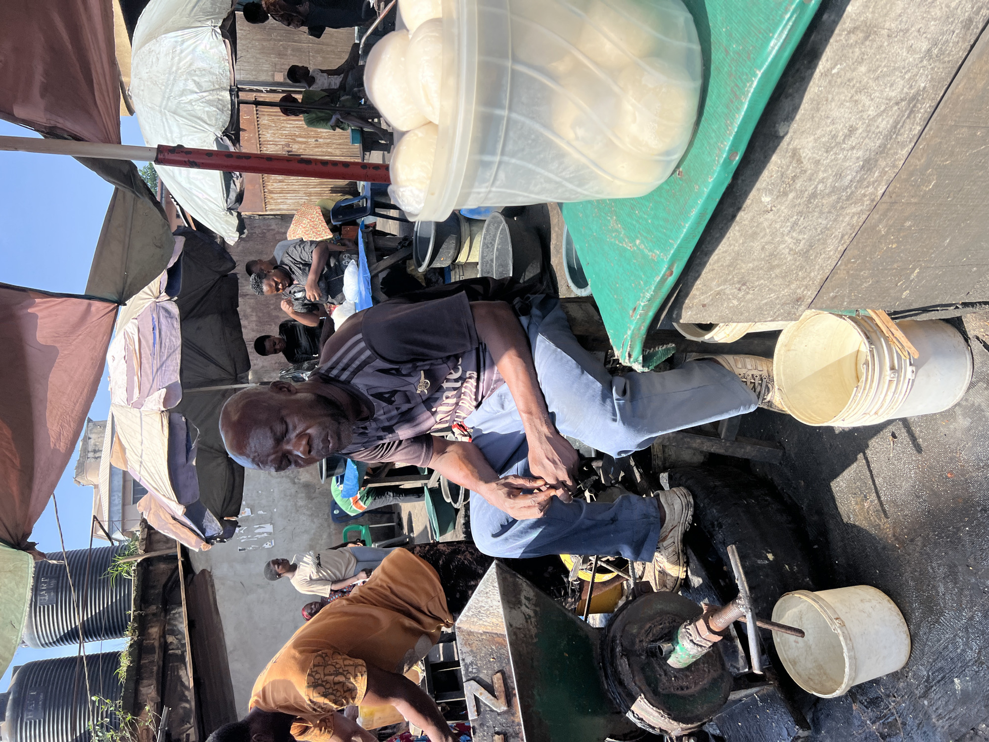 The image captures a vibrant outdoor market scene, likely in Nkpor, Nigeria, during bright daytime. The setting appears informal, characterized by makeshift stalls and shelters providing shade from the clear, sunny sky.
In the foreground, a man with dark skin and a shaved head is seated, wearing a dark grey t-shirt, possibly an Arsenal F.C. jersey, and light blue jeans. He looks downwards, his hands clasped or busy with something in front of him. His expression is neutral and focused. Behind him, other individuals are partially visible, including a person in a black top.
To the upper right, a clear plastic container holds numerous white, spherical food items, possibly fufu balls, resting on a green surface that appears to be part of a market stall or cart. Below this green surface, a dark-colored mechanism with a spout and a handle is visible, accompanied by a white plastic bucket. Another white bucket with a wooden stick inside is on the ground near the man's feet.
Further into the scene, various people are scattered, some seated under tarpaulins and umbrellas of different colors, indicating an active market environment. In the mid-ground, a woman in a light brown/gold patterned top is visible, bending down. In the background, two large, black, ribbed plastic water tanks, one labeled "MART," are stacked vertically, suggesting water storage. A red and white striped pole extends horizontally across the middle of the frame. The ground is a mix of dirt and worn surfaces, typical of a busy outdoor area. The overall atmosphere suggests a typical day of commerce and daily life.