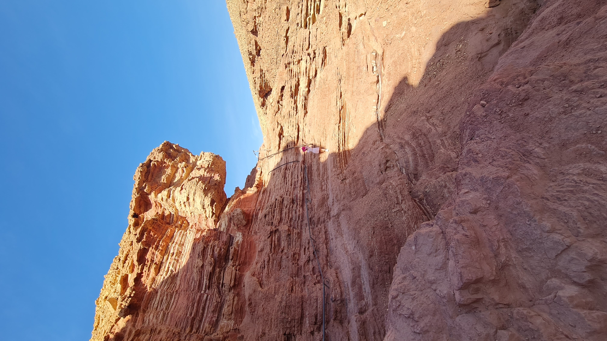 The image shows a person on a via ferrata in a rocky desert environment, possibly in the city of Hevel Eilot Regional Council, Israel. The person is wearing a light-colored outfit and appears to be ascending a cliff face equipped with metal rungs and cables. The rock is predominantly red/brown in color and the sky is clear blue. There is a rock formation on the left and a shadow cast on the cliff face to the right.