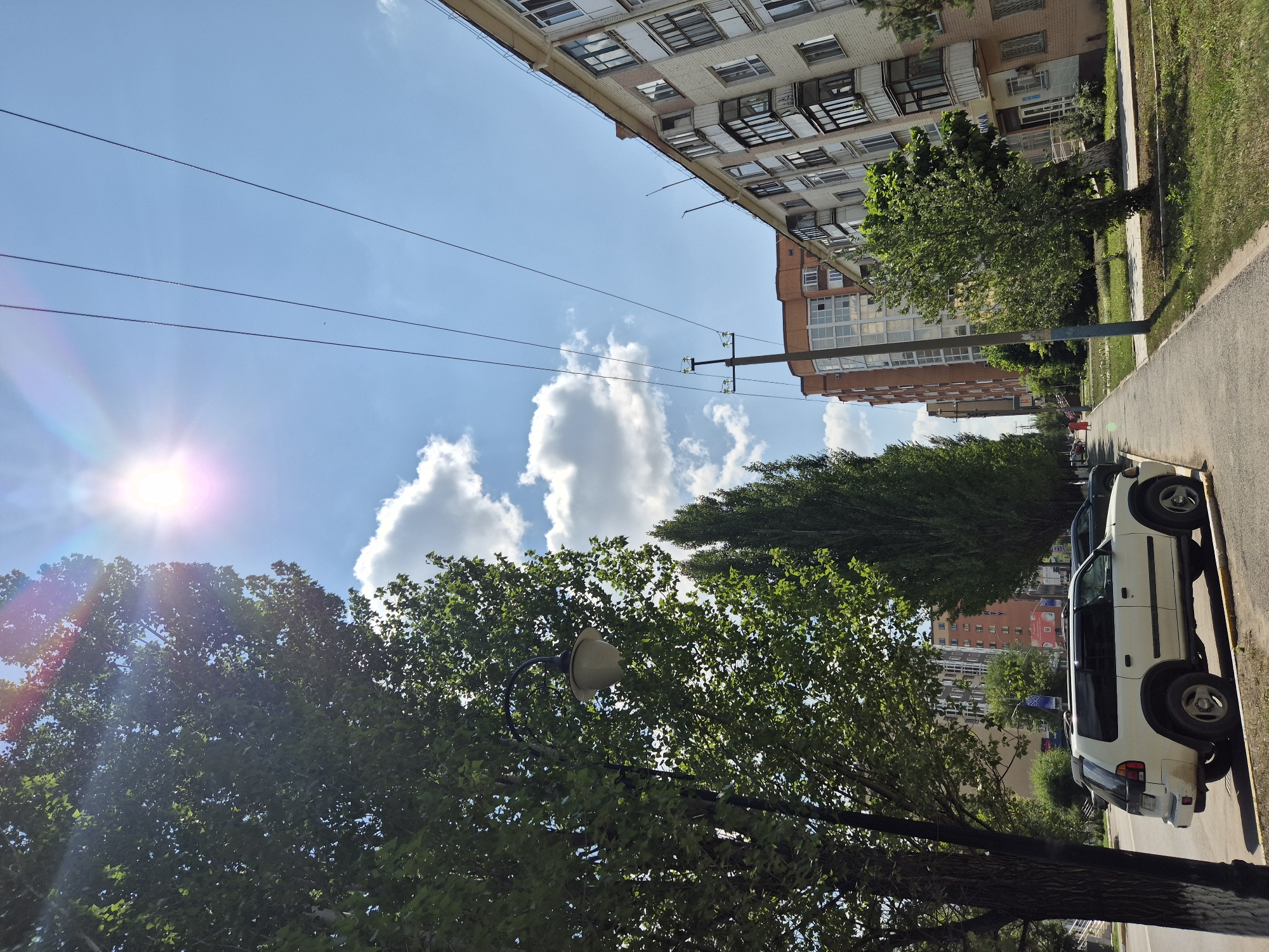 The image captures a sunny day in a residential area of Kokshetau, Kazakhstan. The perspective is from a sidewalk looking down a street lined with trees and apartment buildings. In the foreground, a white SUV is parked on the side of the road, its tires resting on the curb. A street lamp with a light-colored shade is mounted on a pole near the SUV, surrounded by green foliage. Further down the sidewalk, a person in a red outfit is walking away from the camera.
The buildings are a mix of styles, with a light-colored brick apartment building dominating the right side of the frame, featuring balconies and numerous windows. To the left of this building, another taller, red-brick building with many windows is visible. Power lines crisscross the bright blue sky, which is dotted with fluffy white clouds. The sun is high in the sky, casting a bright glare and creating lens flares in the upper portion of the image. Green grass and trees are present along the sidewalk and in front of the buildings, indicating a well-maintained urban environment. The street appears to be paved, with a distinct yellow line marking the edge of the parking area.