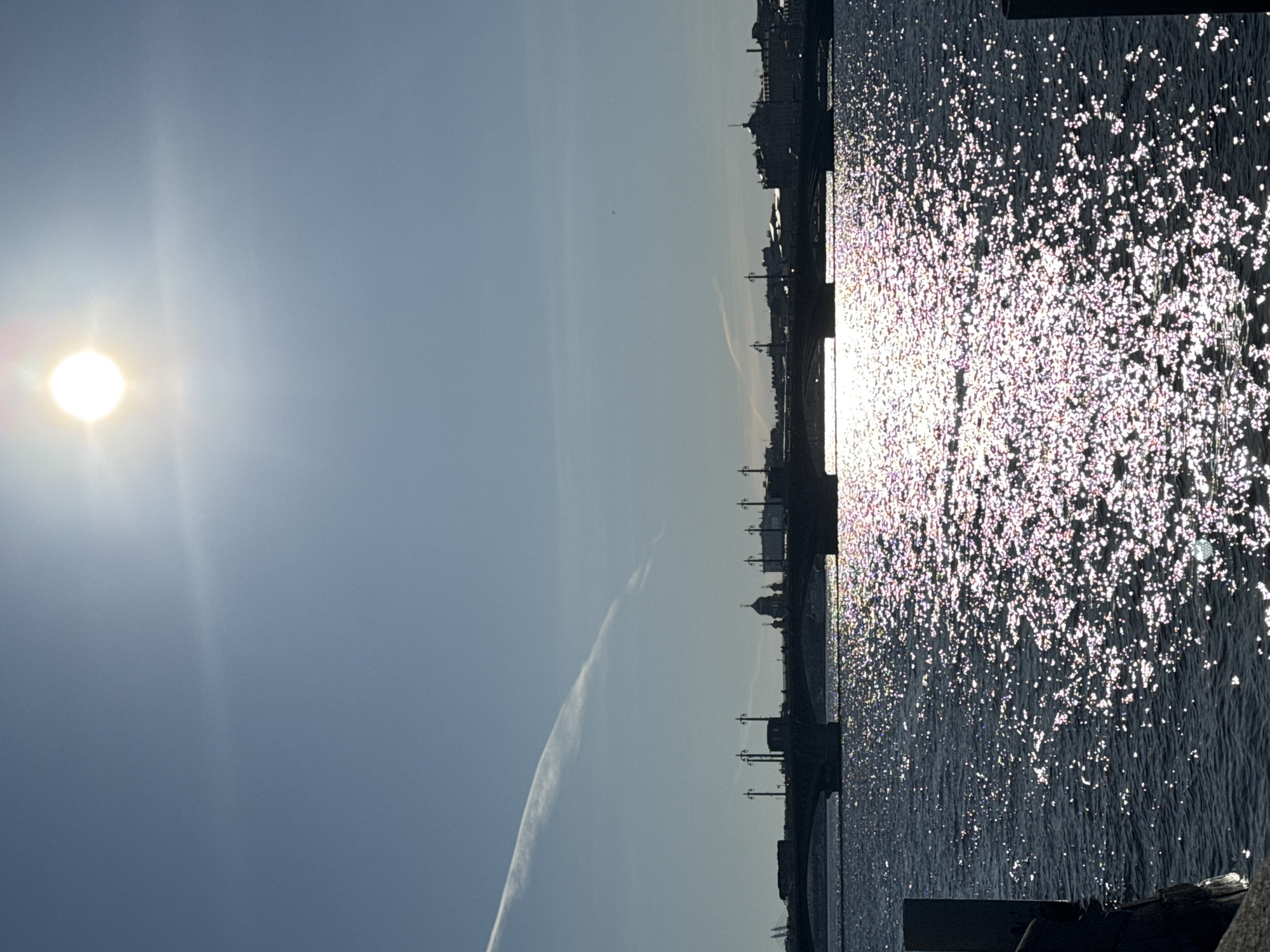 The image depicts a bright, sunny daytime scene of a city skyline across a body of water, likely in Saint Petersburg, Russia, based on the provided context.
The upper left portion of the frame is dominated by a brilliant, circular sun, casting a strong lens flare. The sky is a clear, light blue with a faint, wispy contrail stretching diagonally across the middle.
Along the horizon, silhouetted against the bright sky, lies a distant city. Various buildings, including structures with domes, are visible in dark profile. A bridge, characterized by regularly spaced streetlights, spans across the water, forming part of this silhouette.
The lower right half of the image is filled with the water, which appears dark and rippling. The surface is densely covered with countless sparkling reflections of the sun, creating a dynamic mosaic of bright glints. The strong reflections suggest the sun is at a relatively low angle, indicating either morning or late afternoon. A dark, undefined structure, possibly a quay or boat, is partially visible at the very bottom right edge of the water.
There are no people visible, and no discernible text. The overall atmosphere conveyed is bright, serene, and sparkling.