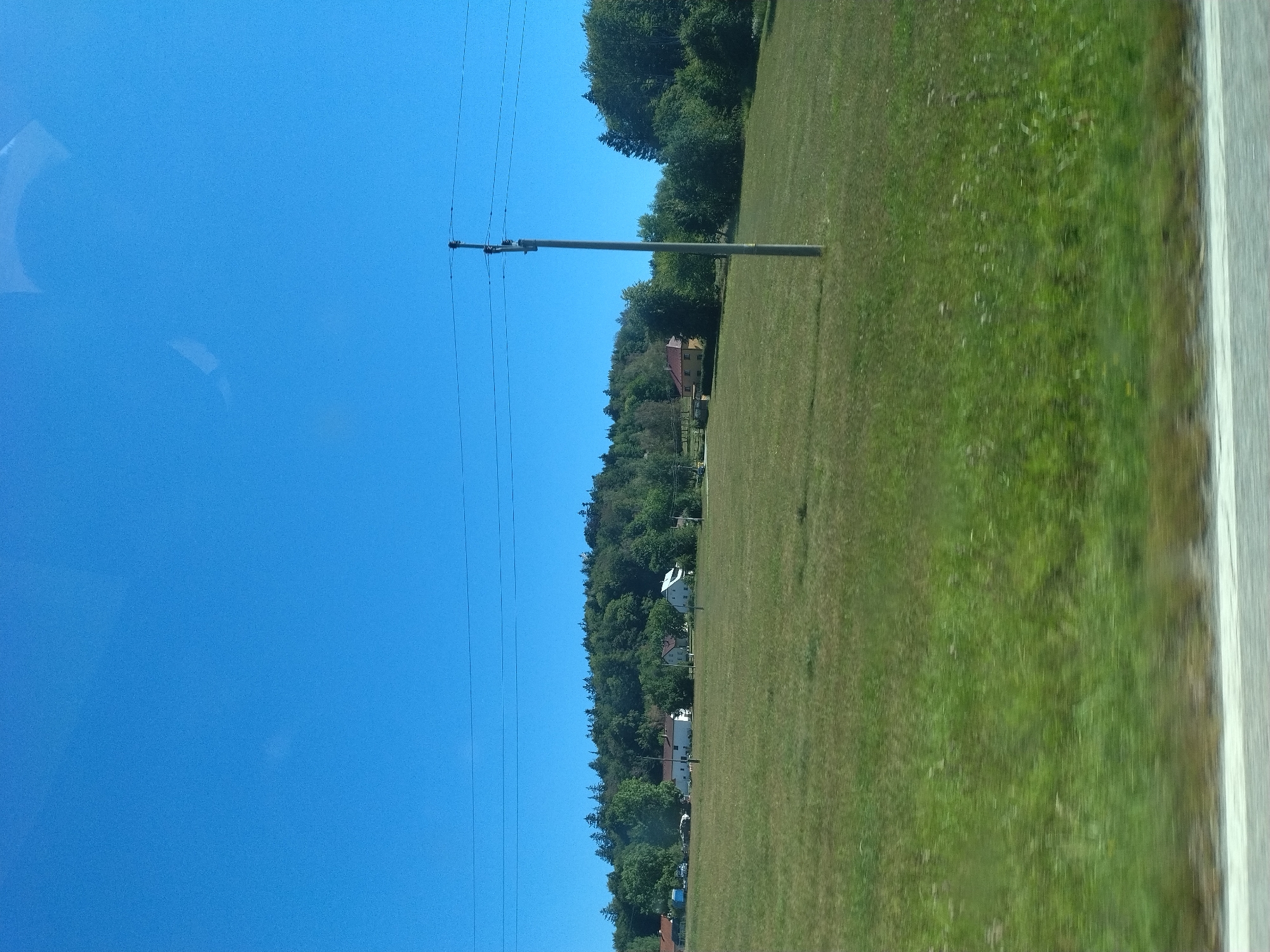 The image depicts a rural landscape with a clear blue sky. In the foreground, a blurred grassy field suggests movement, likely from a vehicle. A paved surface with a white line runs along the very bottom edge. In the mid-ground, several houses are visible, set against a backdrop of a dense, green forest that covers a rolling hill. A solitary utility pole with power lines stands prominently in the field. The overall scene is peaceful and appears to be a sunny day in the countryside. The location is identified as Borová, Czechia.