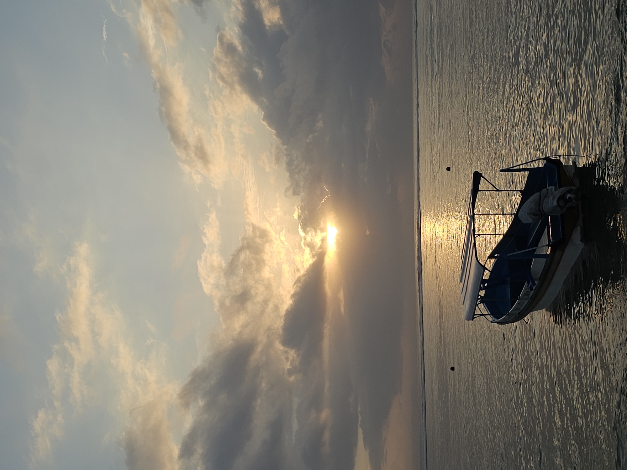 A wide shot captures a serene coastal scene at dusk, likely in Denpasar, Indonesia, based on the presence of a traditional outrigger boat. The sky is dominated by dramatic clouds tinged with the warm hues of the setting sun, which peeks through a break in the clouds, casting a shimmering golden path across the calm ocean. In the foreground, a small, white and blue outrigger boat with a canopy is moored, its reflection visible on the water's surface. Two small buoys are visible in the water, suggesting it's a frequented area. The overall atmosphere is tranquil and picturesque, evoking a sense of peaceful closure to the day.