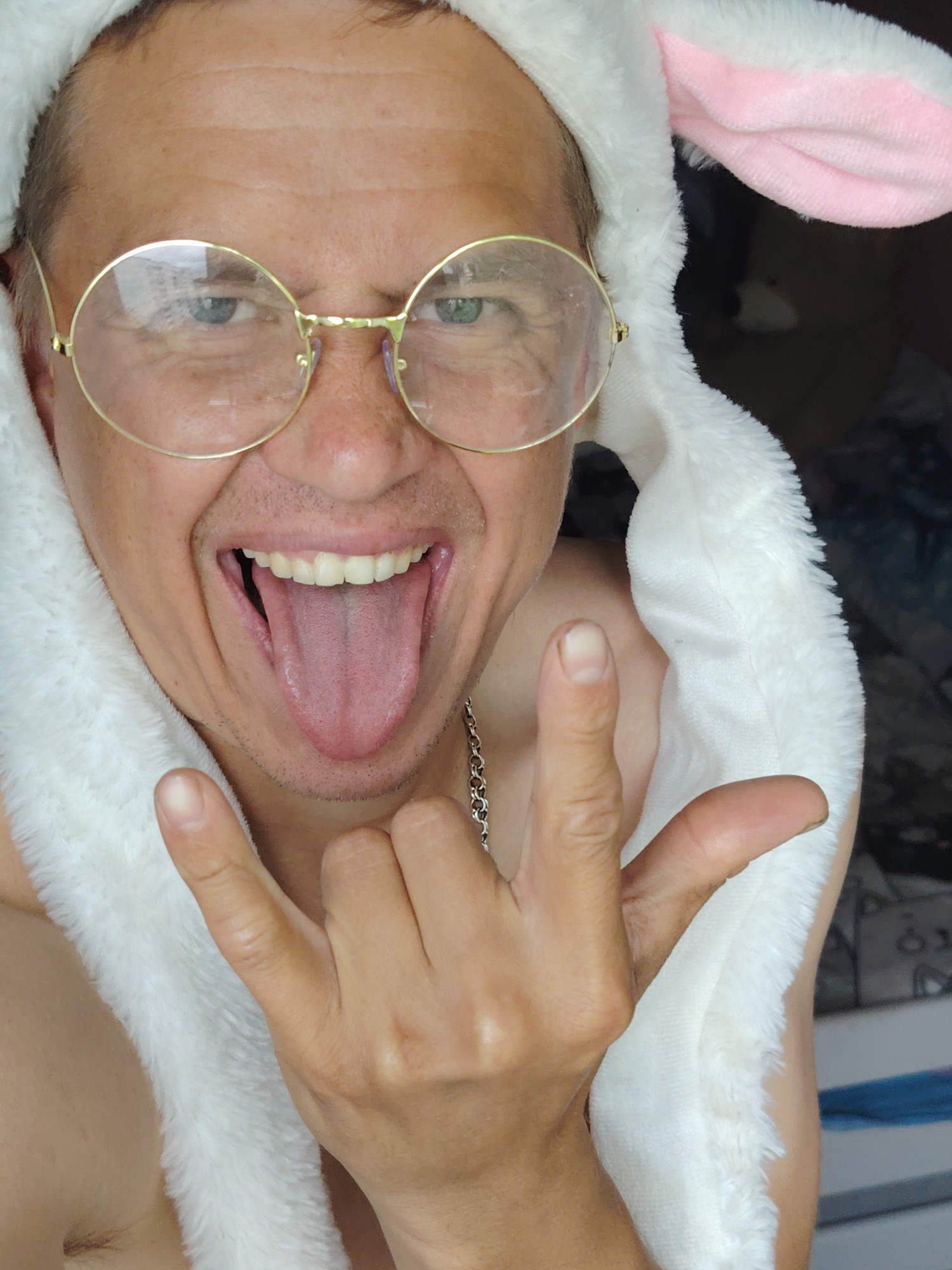 A close-up, high-angle, indoor shot shows a person wearing a white furry hood with pink bunny ears and gold-rimmed glasses. The person is sticking out their tongue, with their mouth wide open. Their right hand is raised, with the index and pinky fingers extended in a "rock on" gesture, while the thumb and middle finger are tucked in. A silver chain necklace is visible around the person's neck. The background is dark and out of focus, with some indistinct objects and a hint of blue and white. The lighting appears to be from above and to the left. The scene is likely from Chaikovsky, Russia, given the context of the prompt.