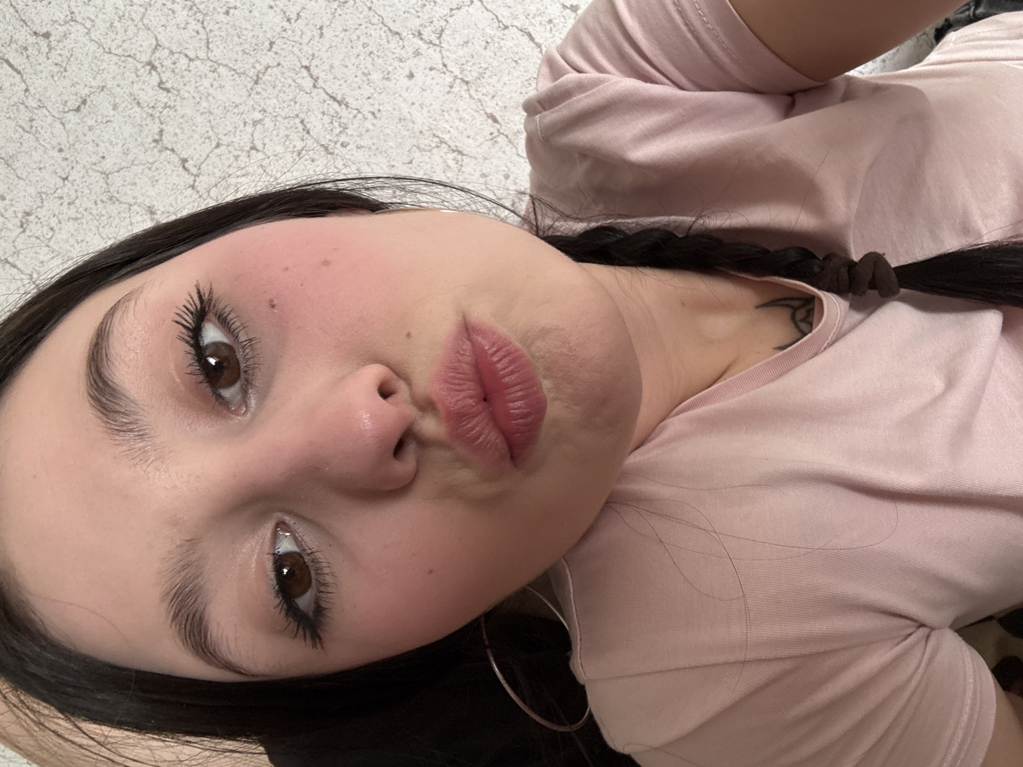 A close-up, angled, selfie-style shot depicts a woman with light skin and dark hair, pursing her lips. Her dark hair is pulled back into a braid secured with a brown hair tie, revealing a tattoo on her neck. She is wearing a light pink t-shirt and a silver hoop earring is visible. The background appears to be a wall with a patterned texture. There is no indication that this image was taken in the city of Фоки, Russia.