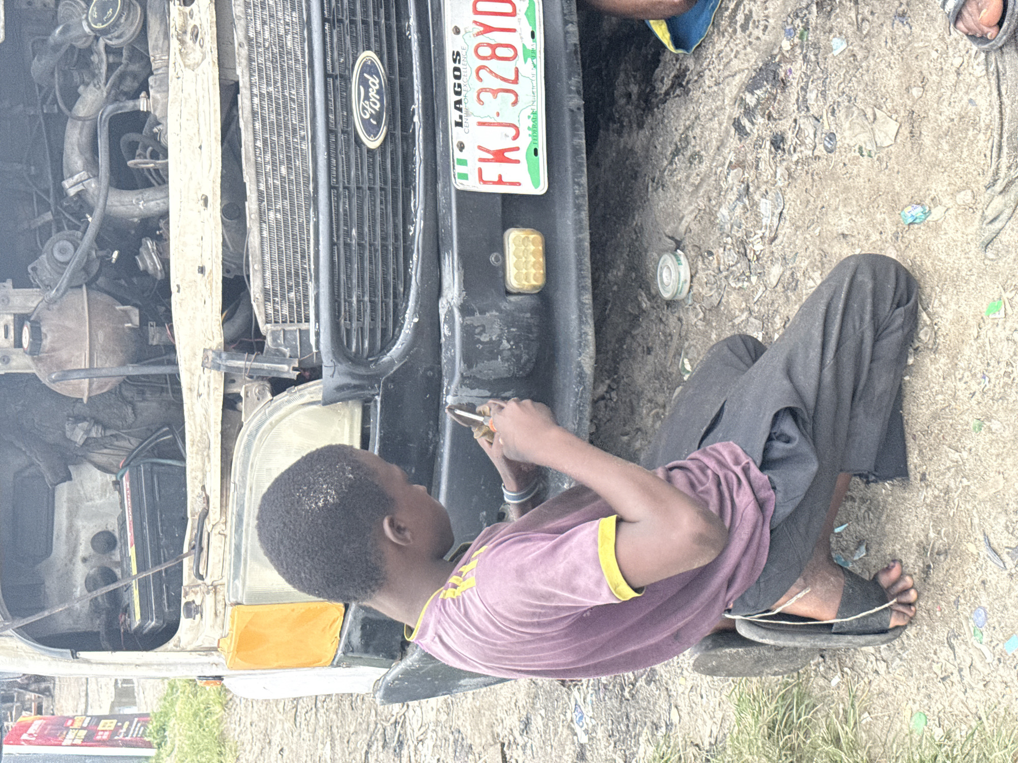 A young boy in a purple t-shirt with yellow stripes on the sleeves and dark pants kneels on the ground, facing a disheveled white Ford van. The van's hood is open, revealing a complex engine compartment. The boy appears to be working on the front of the van, holding a small tool in his right hand and a smaller item in his left. A Lagos, Nigeria license plate, FKJ-328YD, is affixed to the front bumper. The ground is rough and appears to be a mix of dirt and debris, with some green grass visible on the left side of the frame. The overall impression is that of a roadside repair scene in a somewhat unkempt urban environment in Ado, Nigeria.